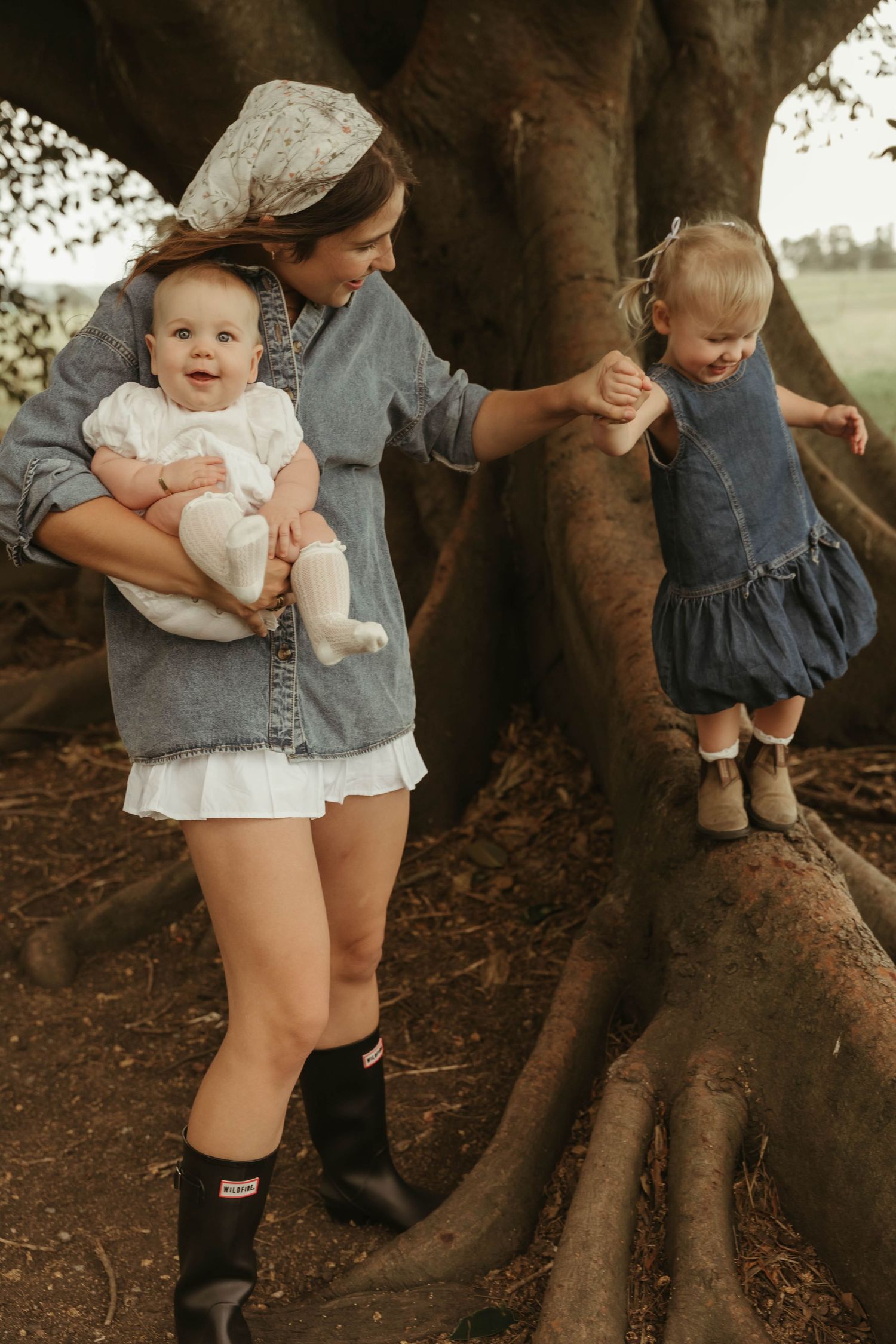 Mother holds infant while helping toddler climb a large tree in a park setting.
