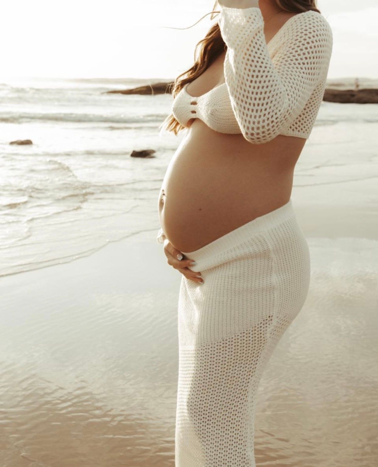 Pregnant woman in white crochet outfit cradling baby bump on a serene beach with ocean waves.