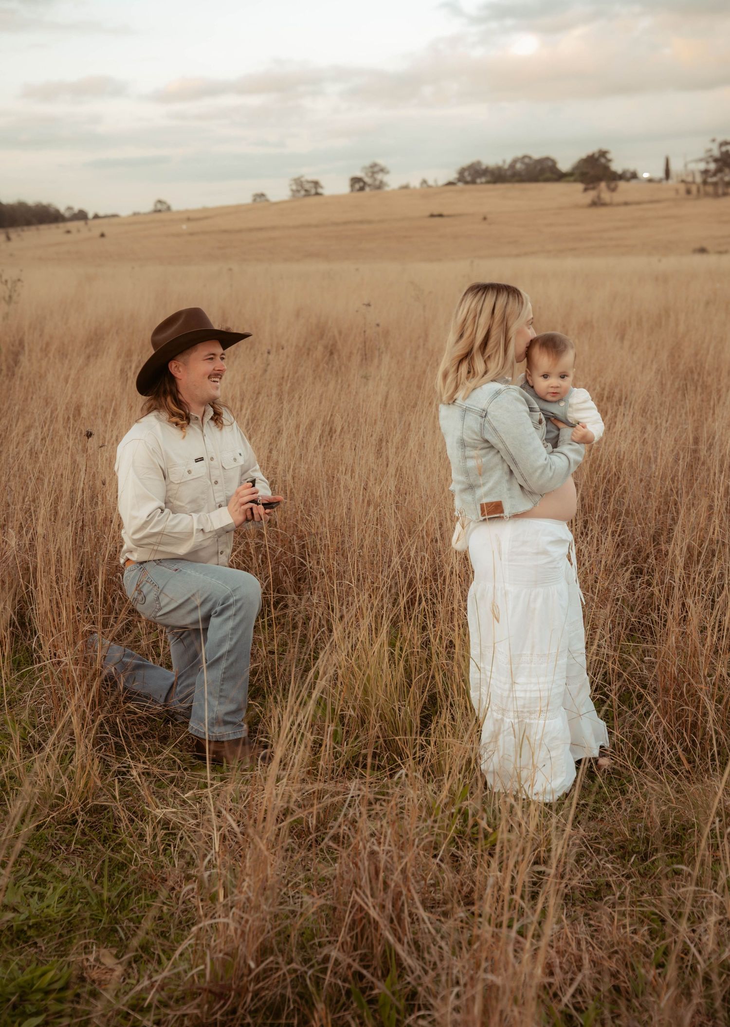 Man kneeling in golden field proposing to woman holding baby, rustic countryside engagement photo at dusk.