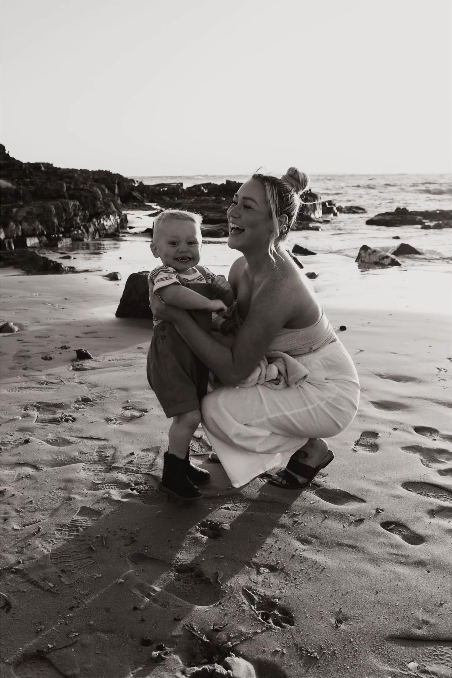 Black and white photo of a smiling mother crouching with her toddler on a sandy beach.