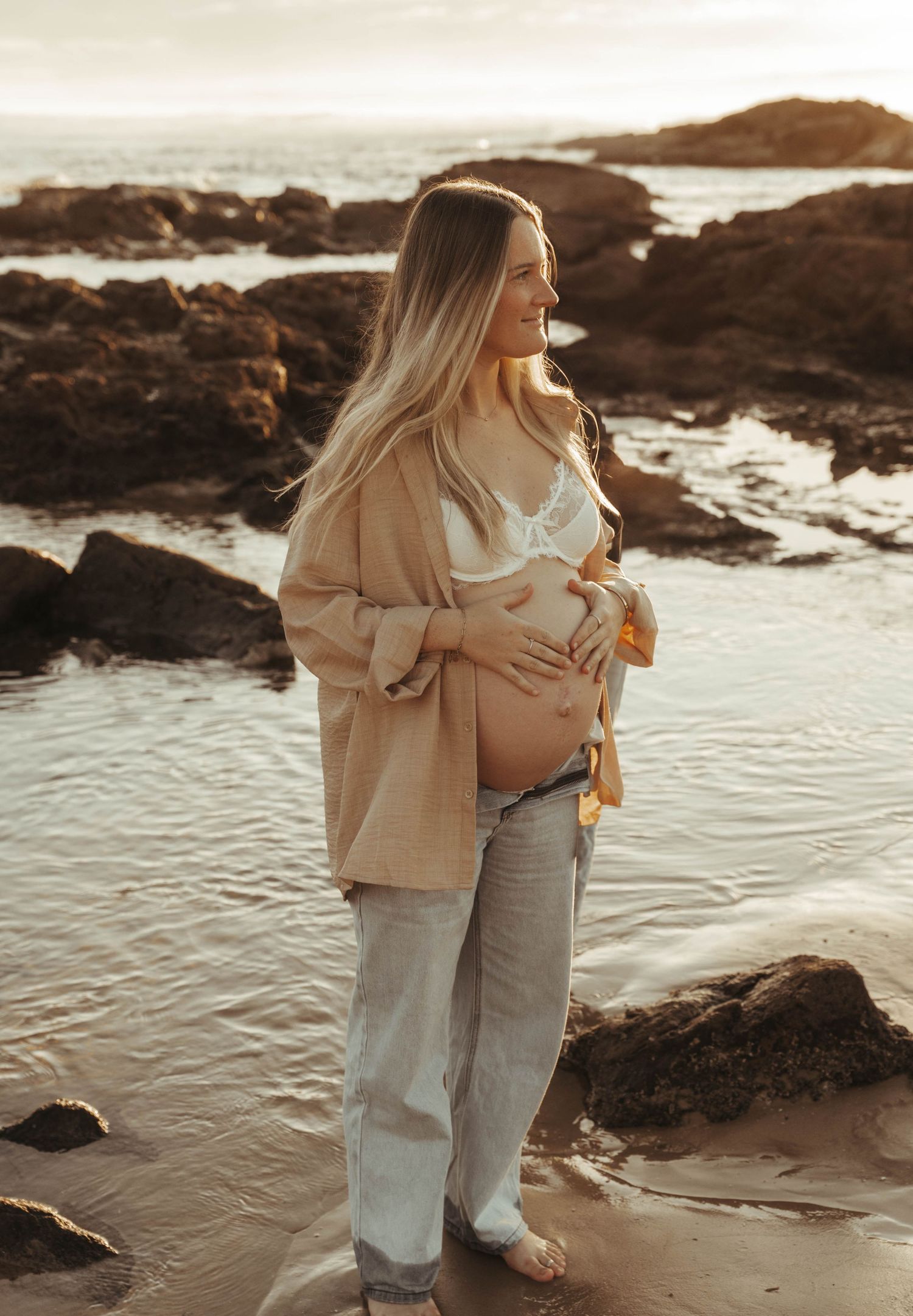 A pregnant woman cradles her baby bump on a rocky beach at sunset in a beige blazer and jeans.