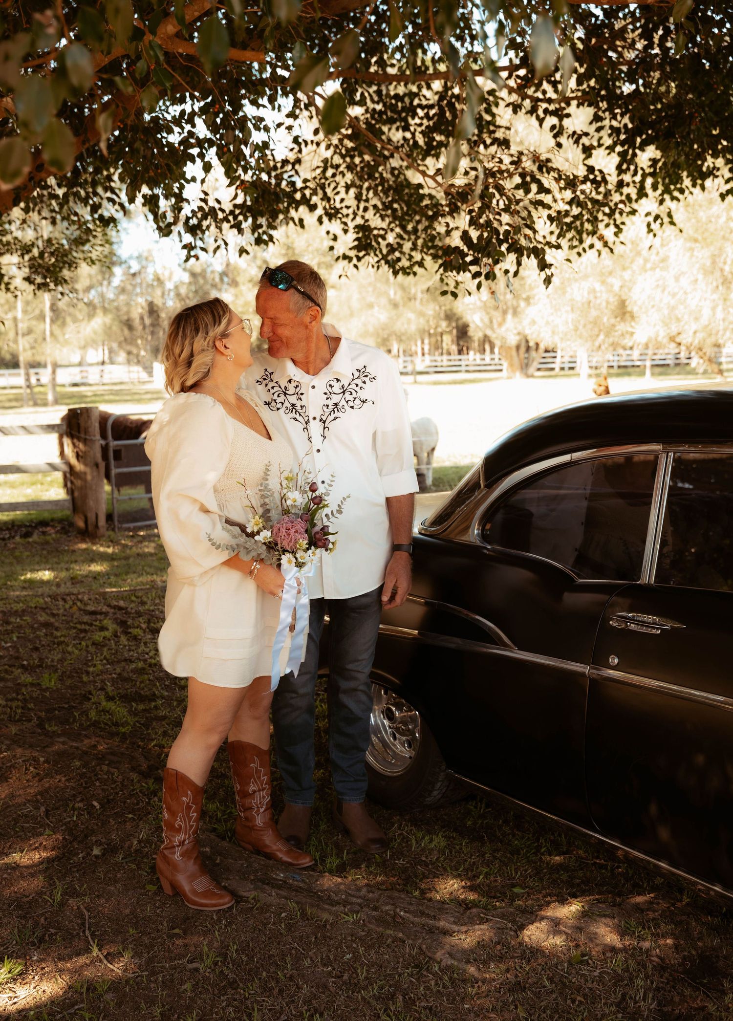 Couple sharing a romantic moment beside a vintage black car on a rustic farm, bride in white dress and cowboy boots holding wildflowers.