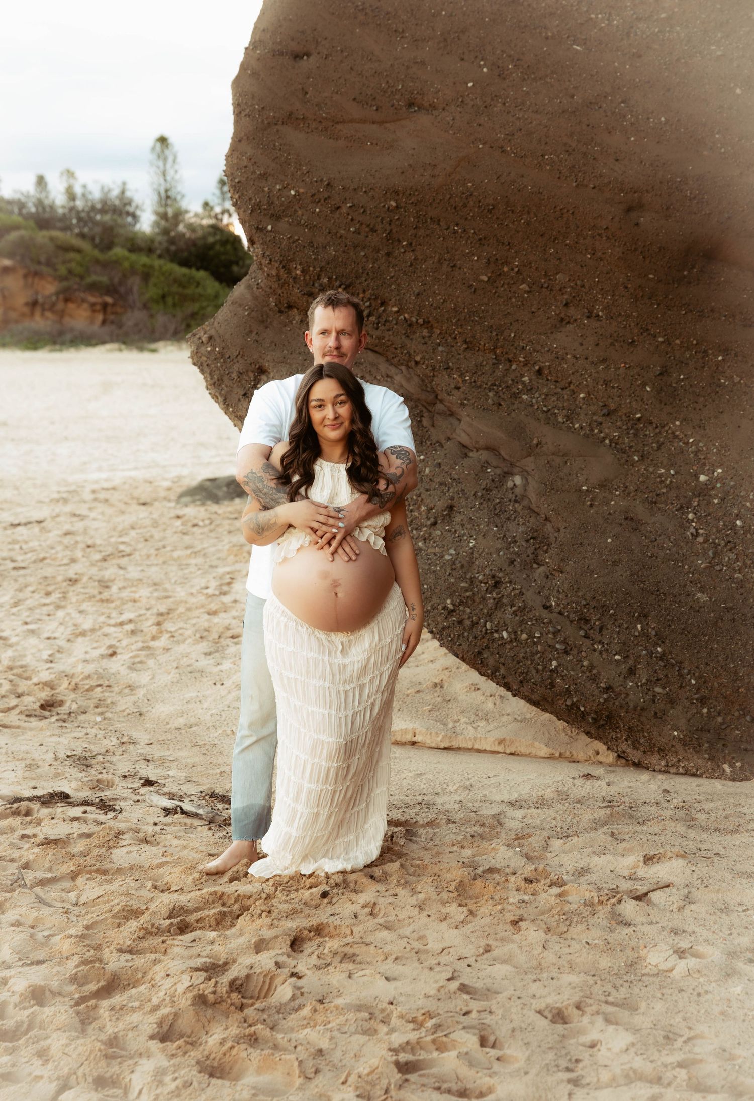 Couple posing on sandy beach by large rock during maternity photoshoot, woman in white lace dress showing baby bump.