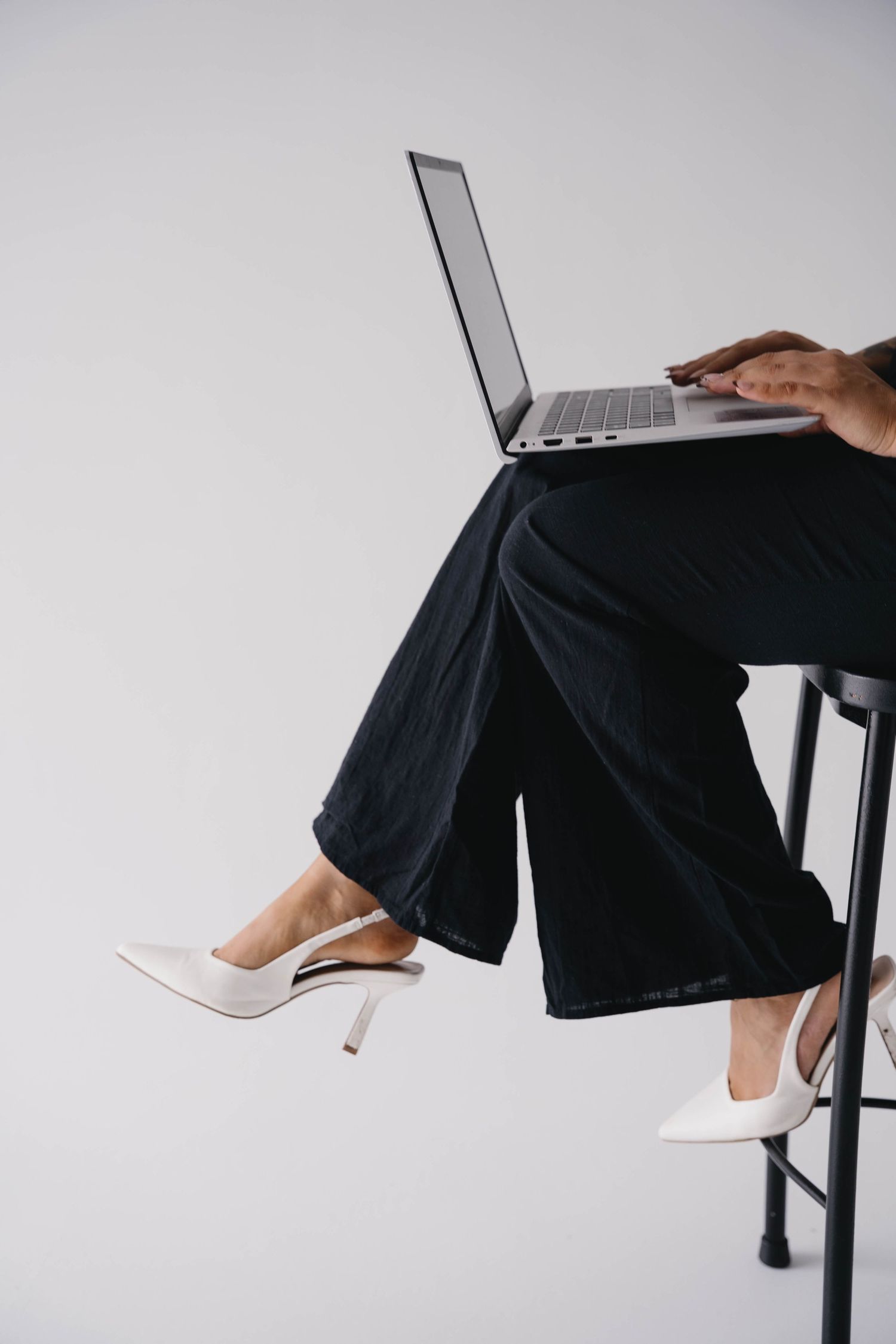 Woman in black wide-leg pants and white heels sitting at a desk working on a laptop against a white background.
