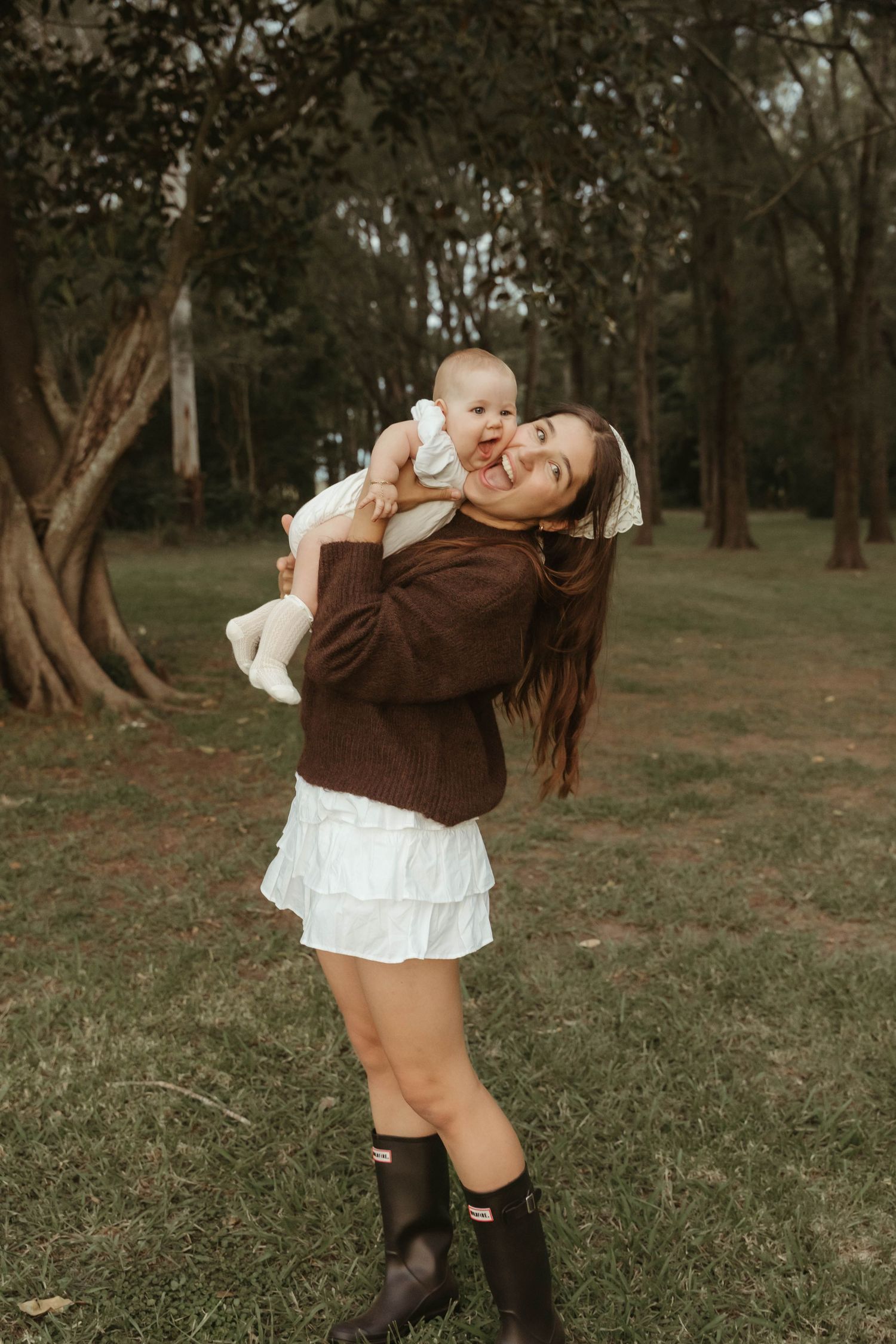 Smiling mother lifting happy baby overhead in a lush green park, wearing brown sweater and white skirt.