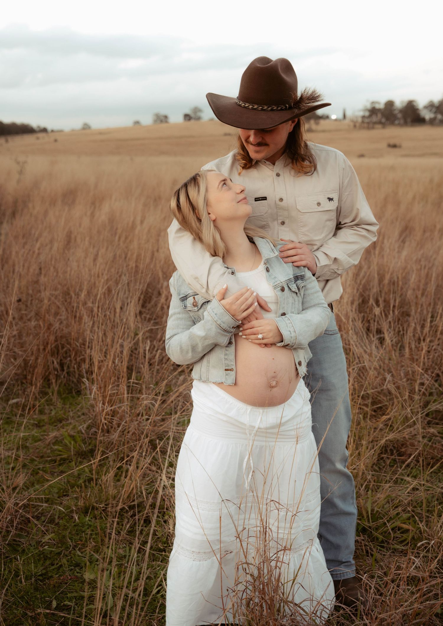Couple sharing tender maternity moment in golden wheat field, man in cowboy hat embracing pregnant partner.