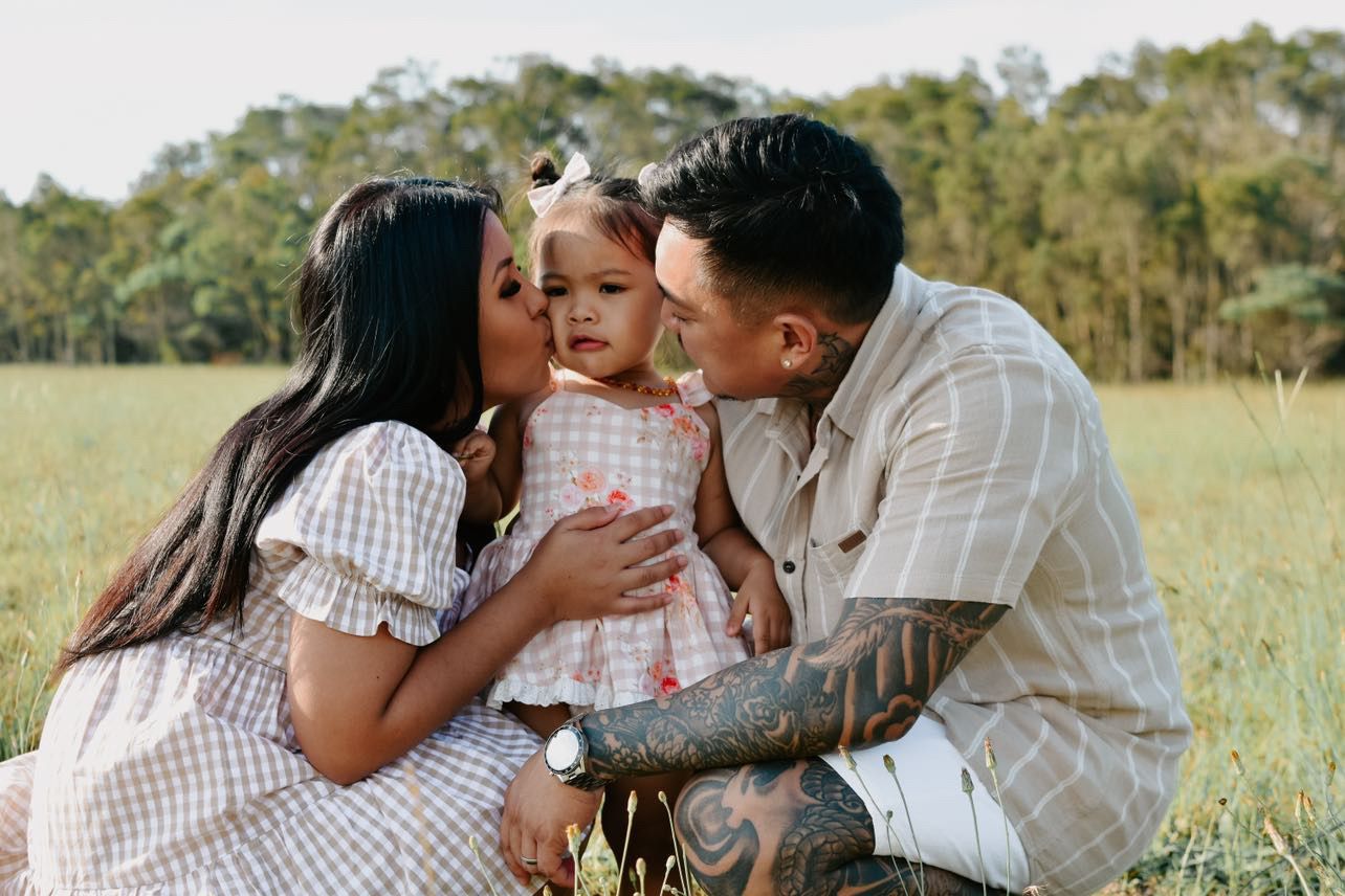 A couple kisses their toddler daughter on the cheeks during a family photo session in a green field.