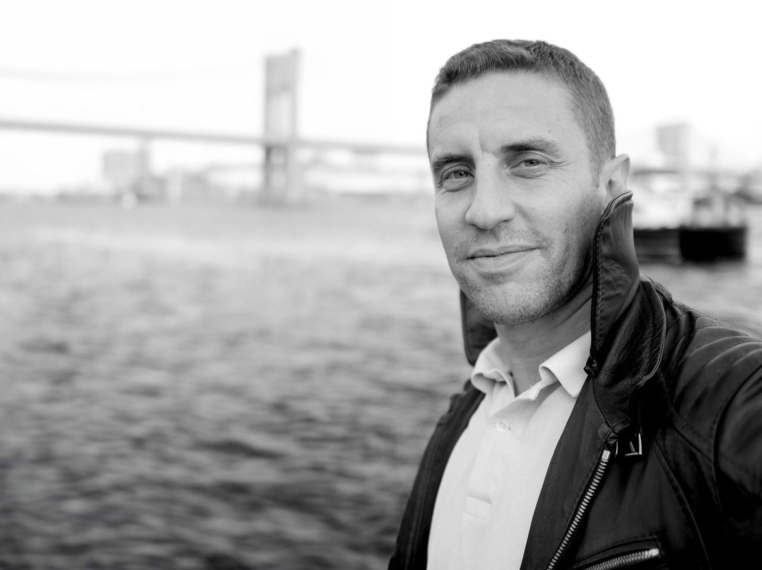 Black and white portrait of a man smiling near a waterfront with a bridge in the background.