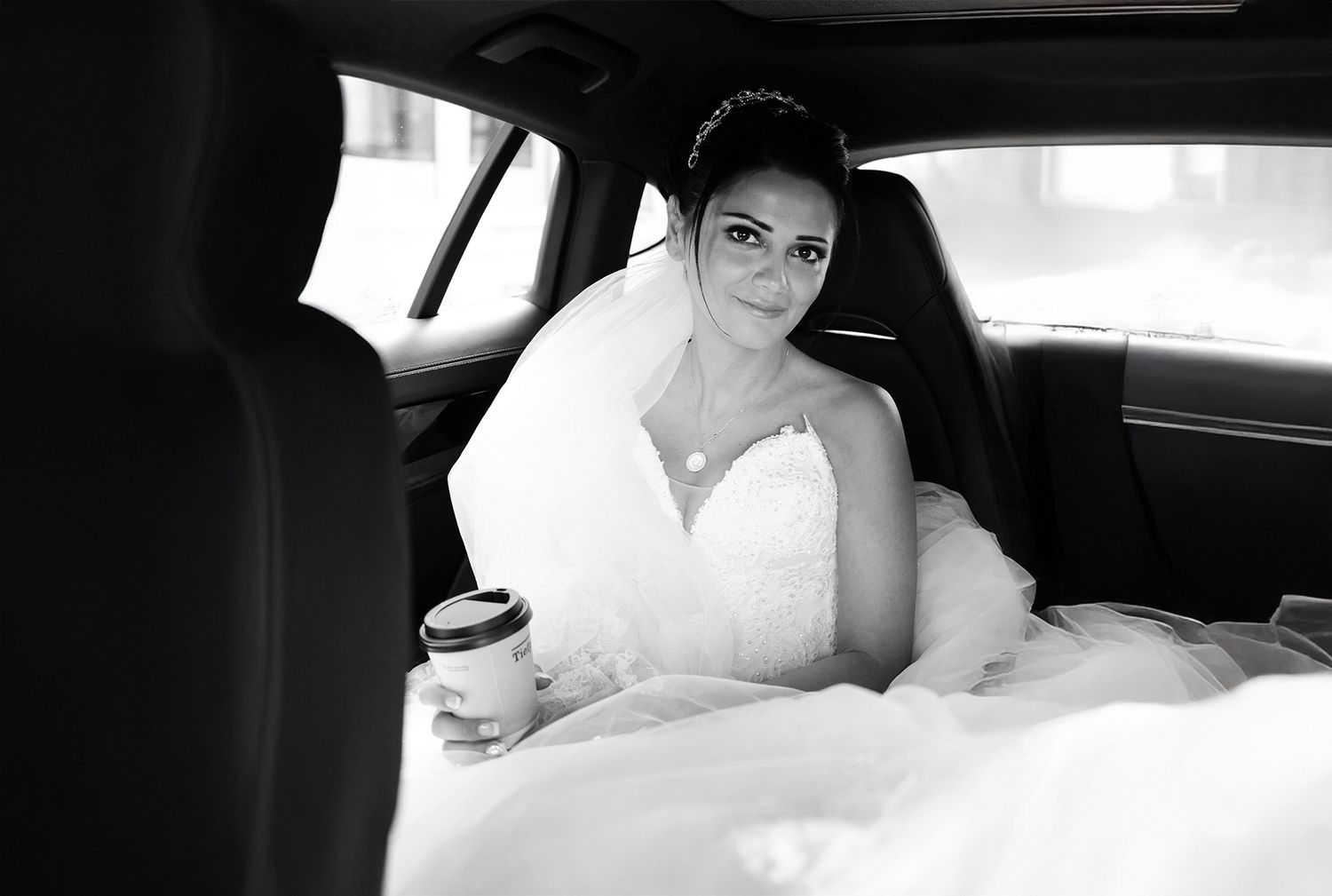 Black and white photo of a smiling bride in a wedding dress holding a coffee cup in a car.