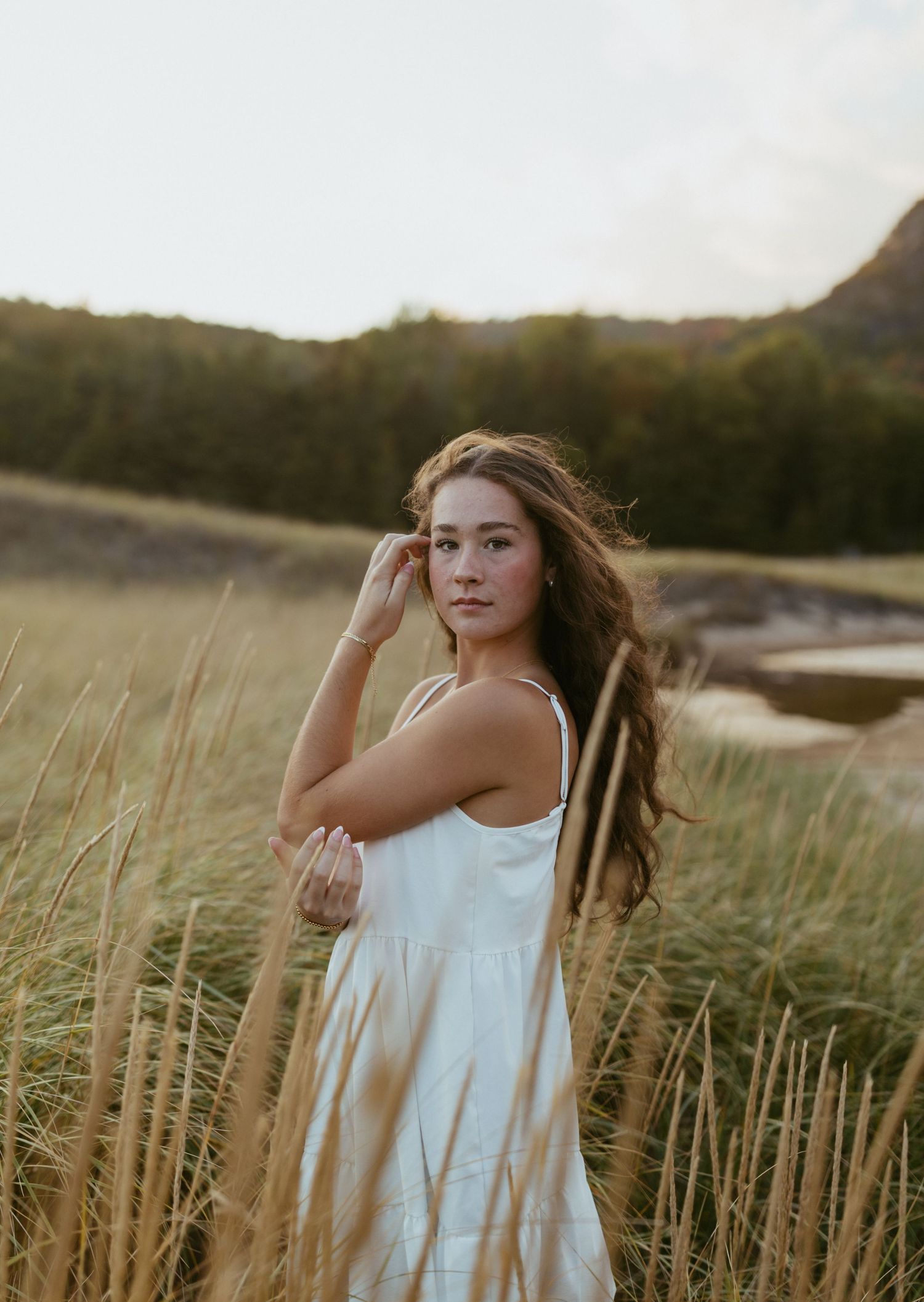Young woman in white sundress standing in golden tall grass field at sunset with mountains behind her.
