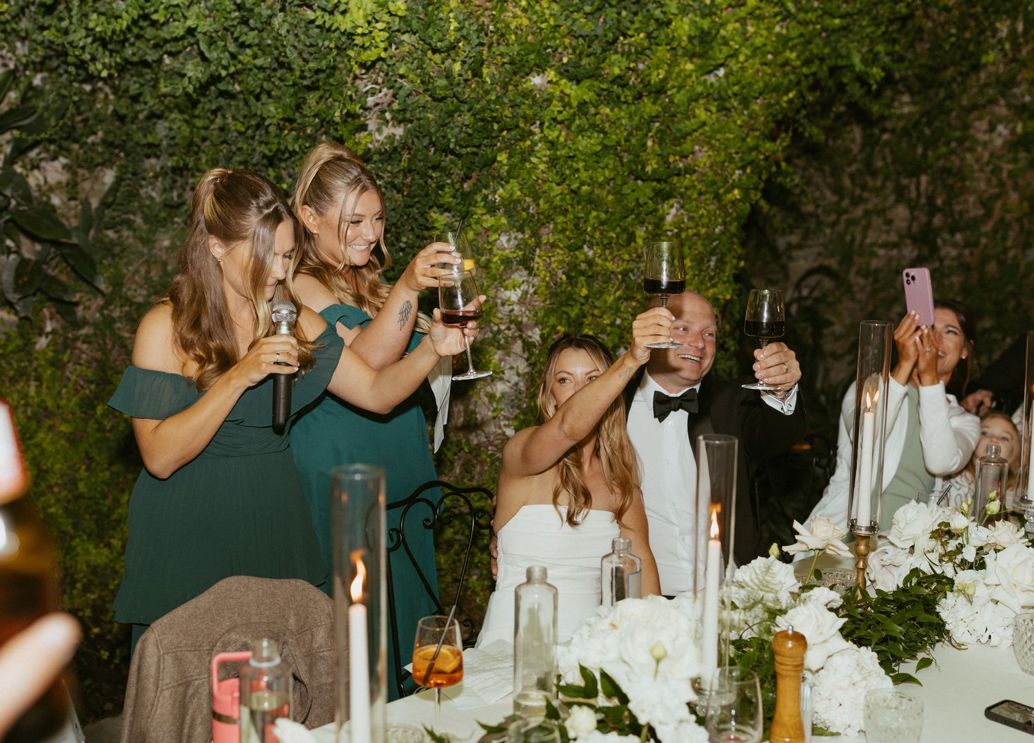 Wedding guests in green dresses raise champagne glasses in a toast at an elegant reception with white floral arrangements.