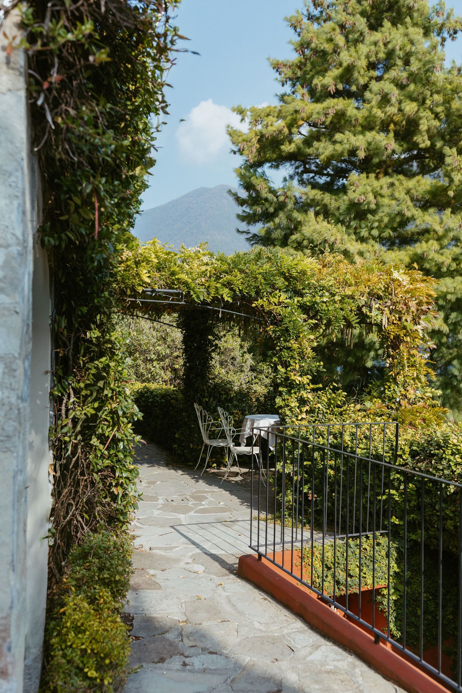 A lush ivy-covered garden pathway with stone tiles, metal railing, and chairs overlooking a mountain view.