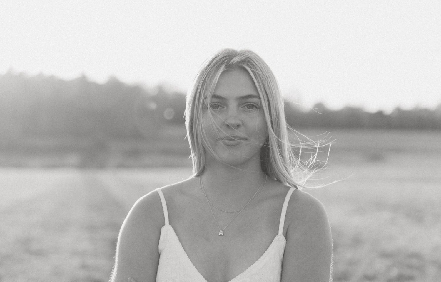 Black and white portrait of a young blonde woman in a white dress standing in an open field.