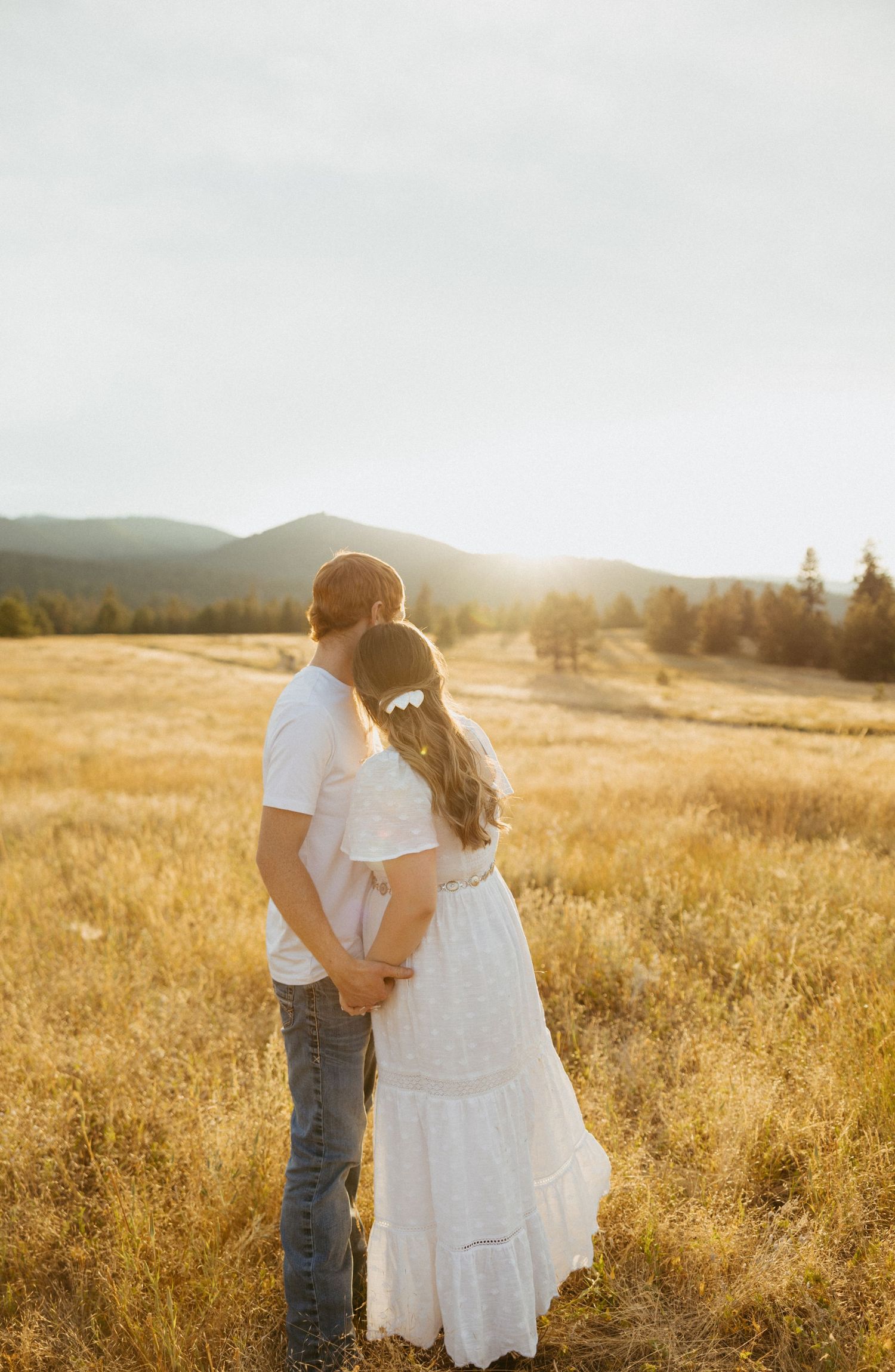 Couple holding hands in golden field at sunset, woman in white dress with flower hair accessory, mountains in background.