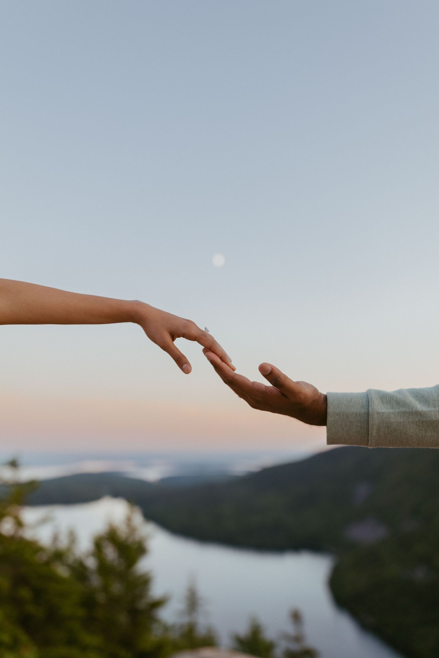 Two hands reaching toward each other with a moonlit mountain river valley below.