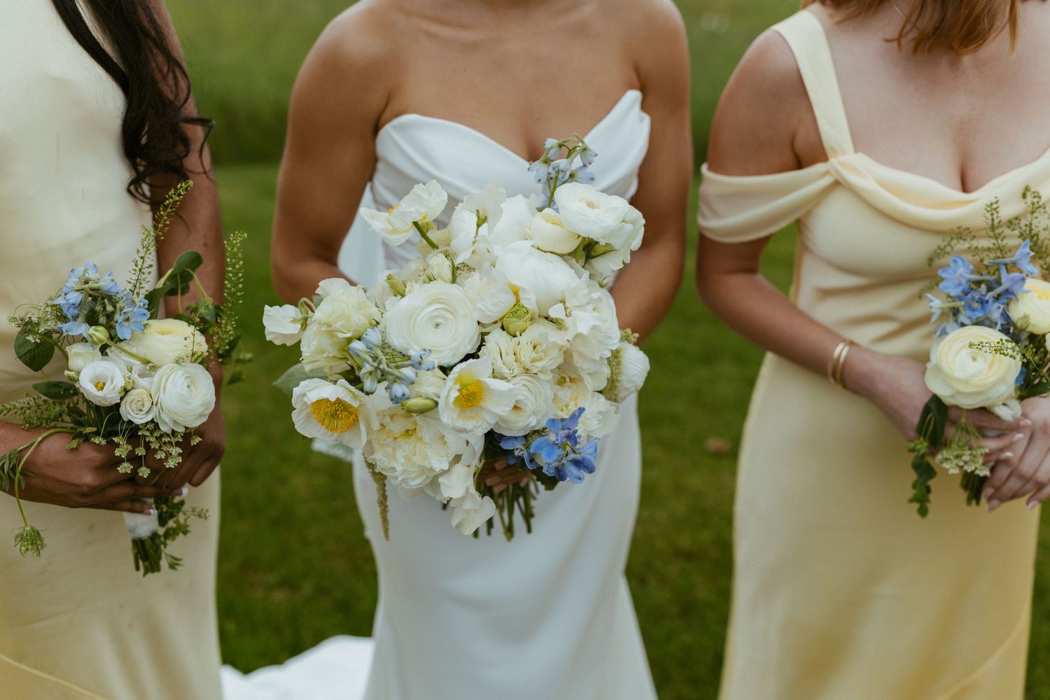 Bride in white strapless gown with white and blue bouquet flanked by bridesmaids in yellow dresses at outdoor wedding.