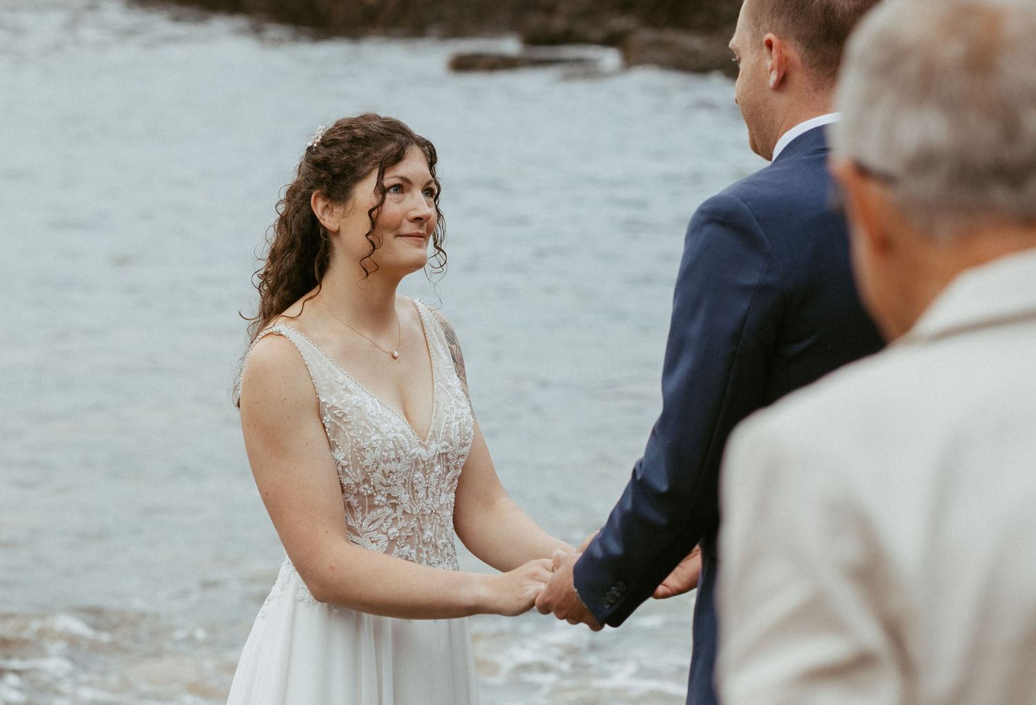 Bride smiling joyfully while holding hands with groom during an outdoor beach wedding ceremony.