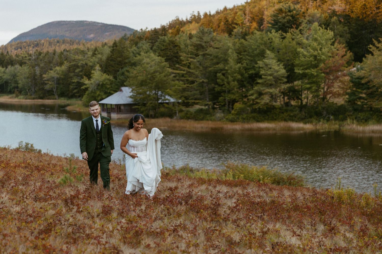 Newlyweds walk hand in hand through an autumn meadow with colorful foliage and a lake.