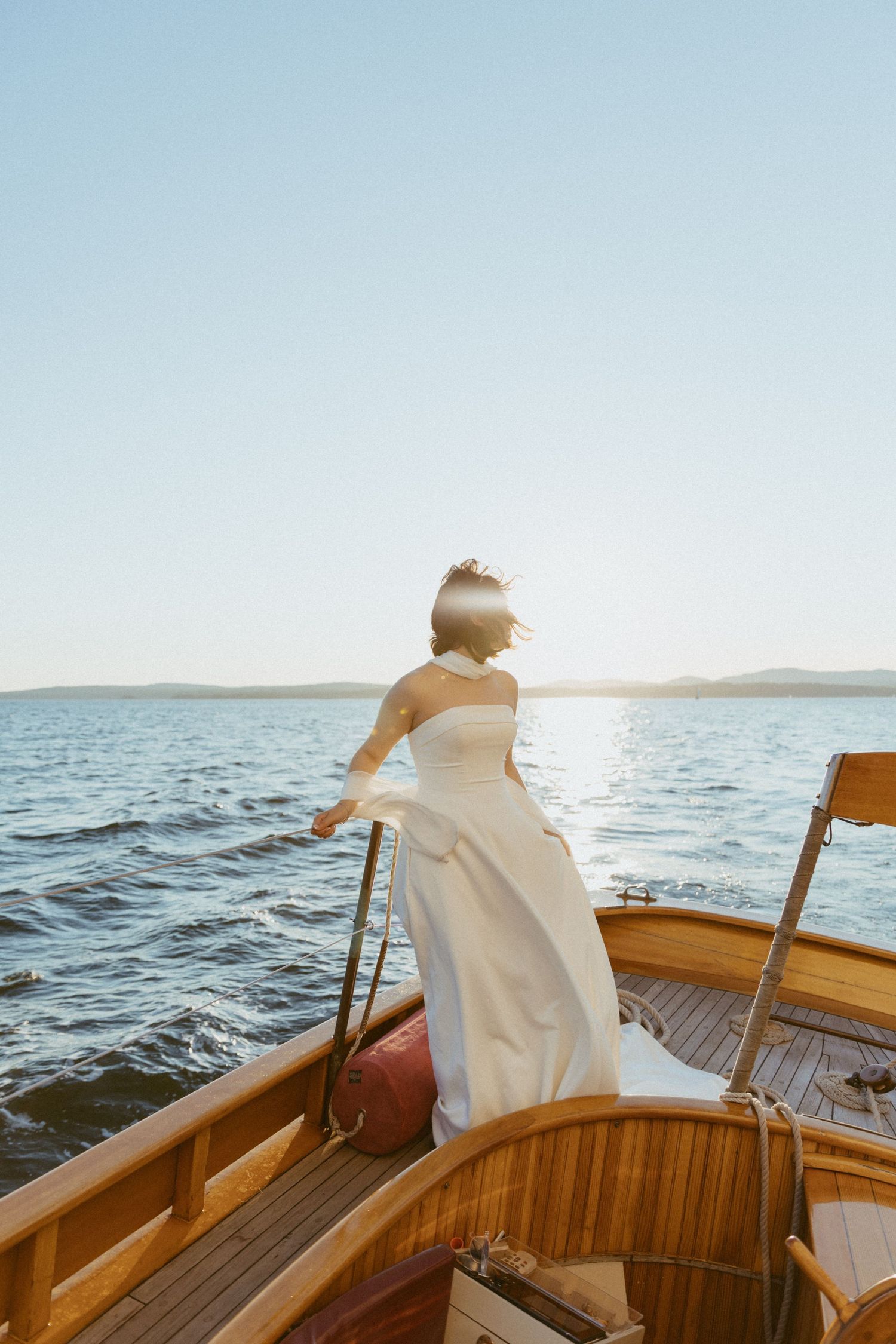 Bride in elegant white dress standing at bow of wooden sailboat overlooking calm ocean at sunset.
