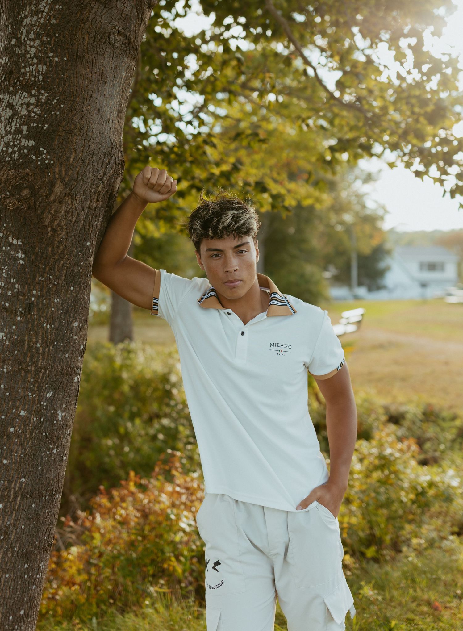 Young man in white polo shirt and shorts leaning against a tree in a sunny outdoor setting.