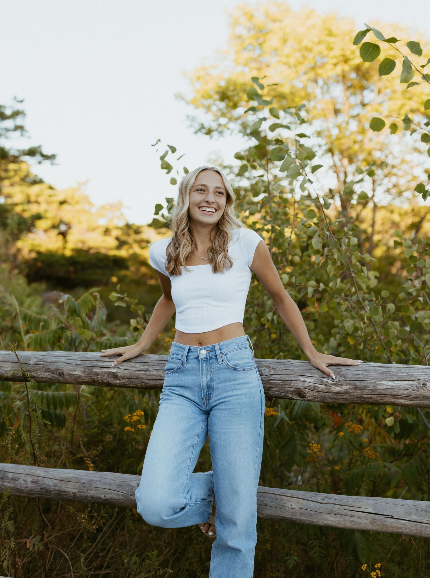 Happy blonde woman in white crop top and wide-leg jeans leaning on a rustic wooden fence outdoors.