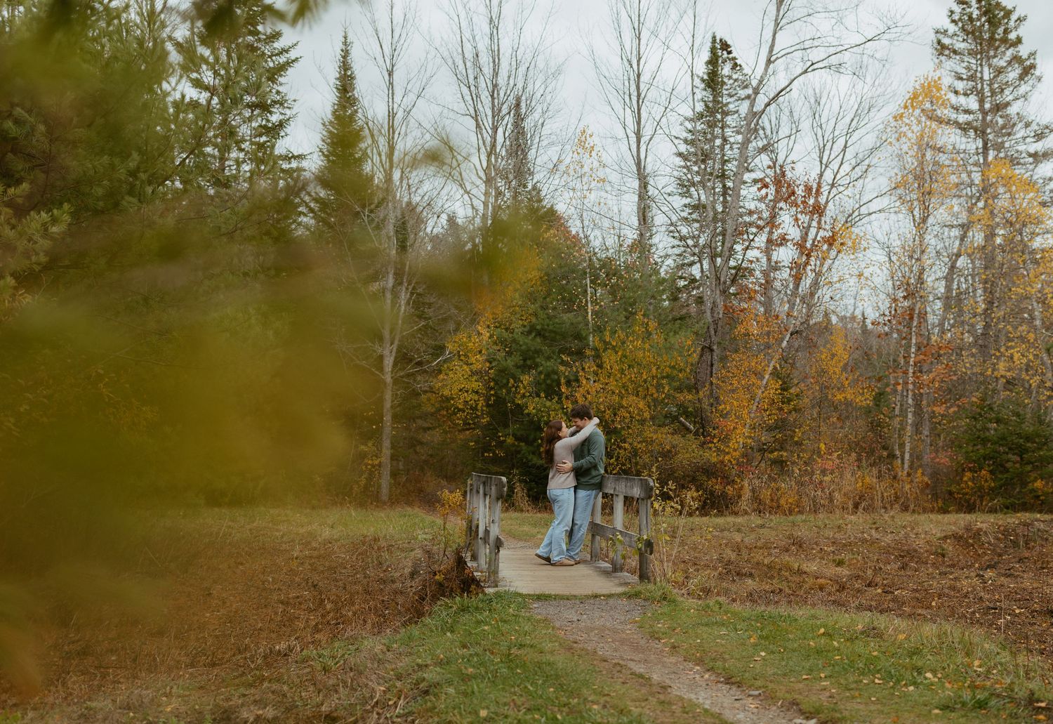 Couple embracing on a wooden bridge path surrounded by colorful fall trees and foliage.