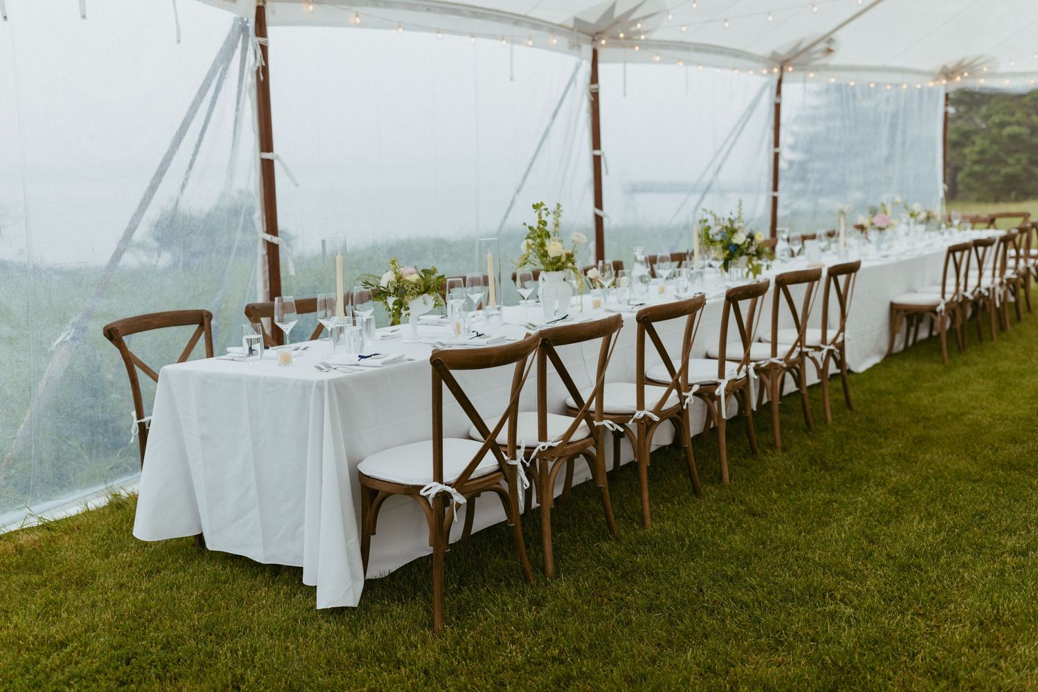 Elegant outdoor wedding reception setup with long white-clothed table, wooden chairs under clear tent on grassy lawn.