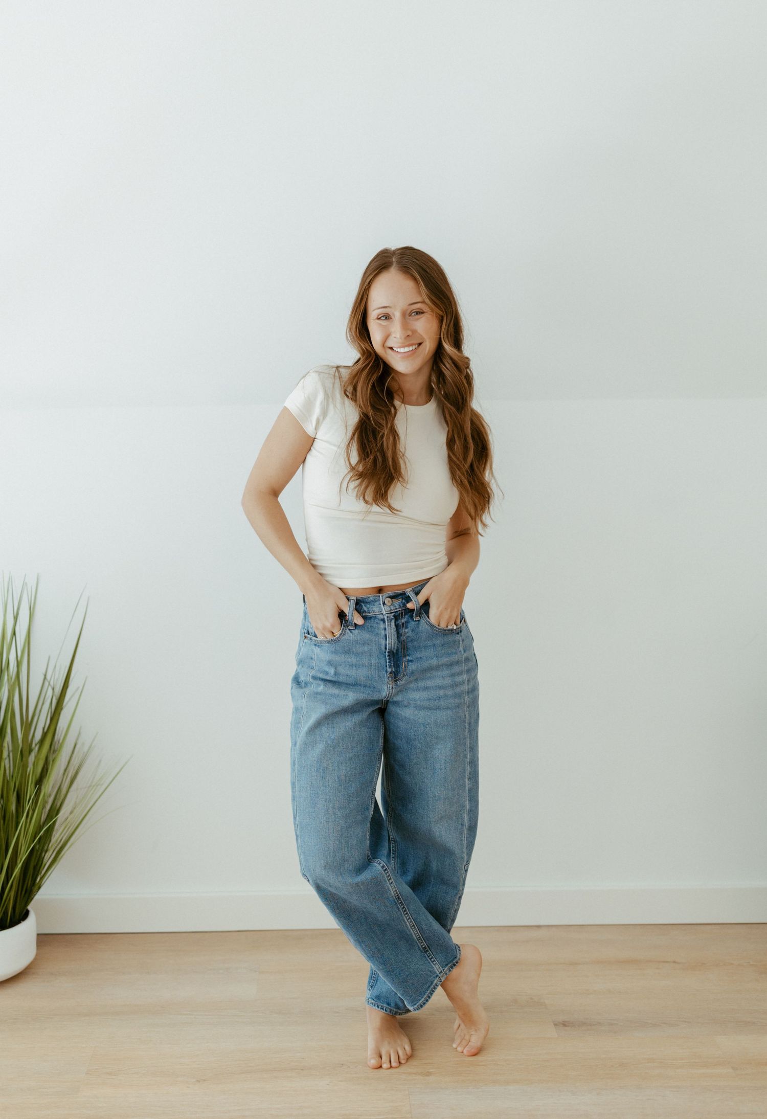 Smiling woman in white crop top and wide-leg jeans poses barefoot against a light wall with hands in pockets.