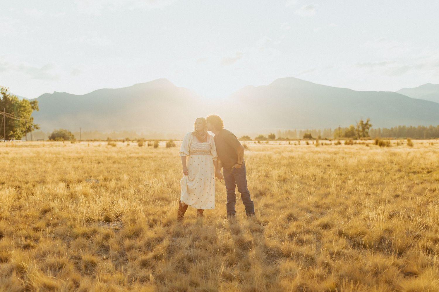 Couple sharing a romantic kiss in a golden sunlit field with mountain backdrop at sunset.