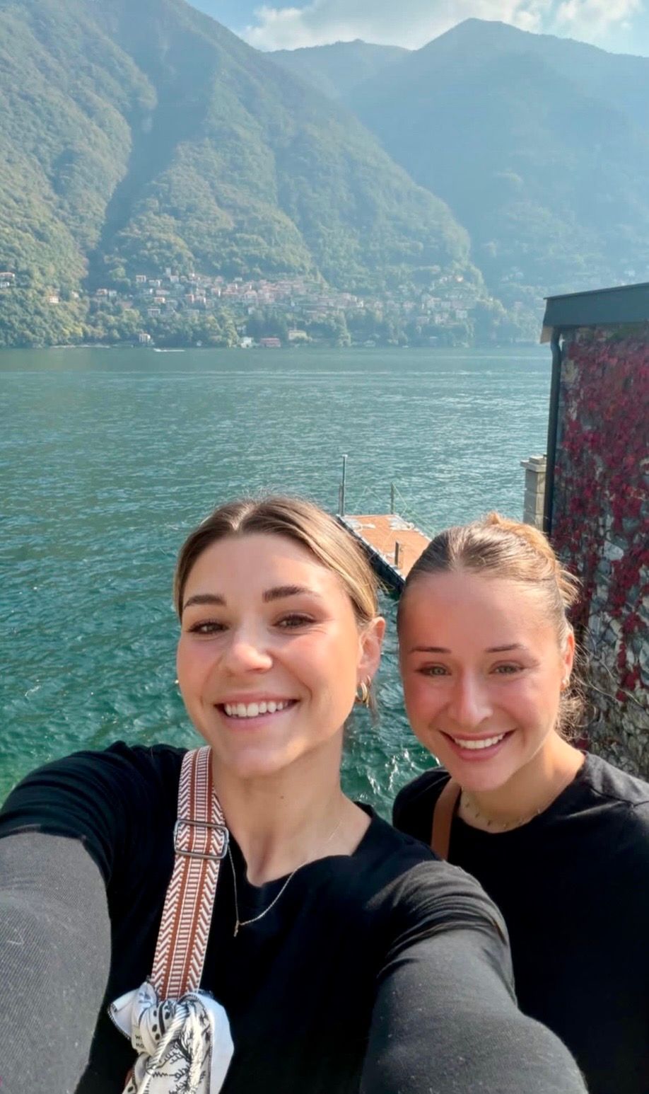 Two smiling women take a selfie by a turquoise alpine lake with green mountains in the background.