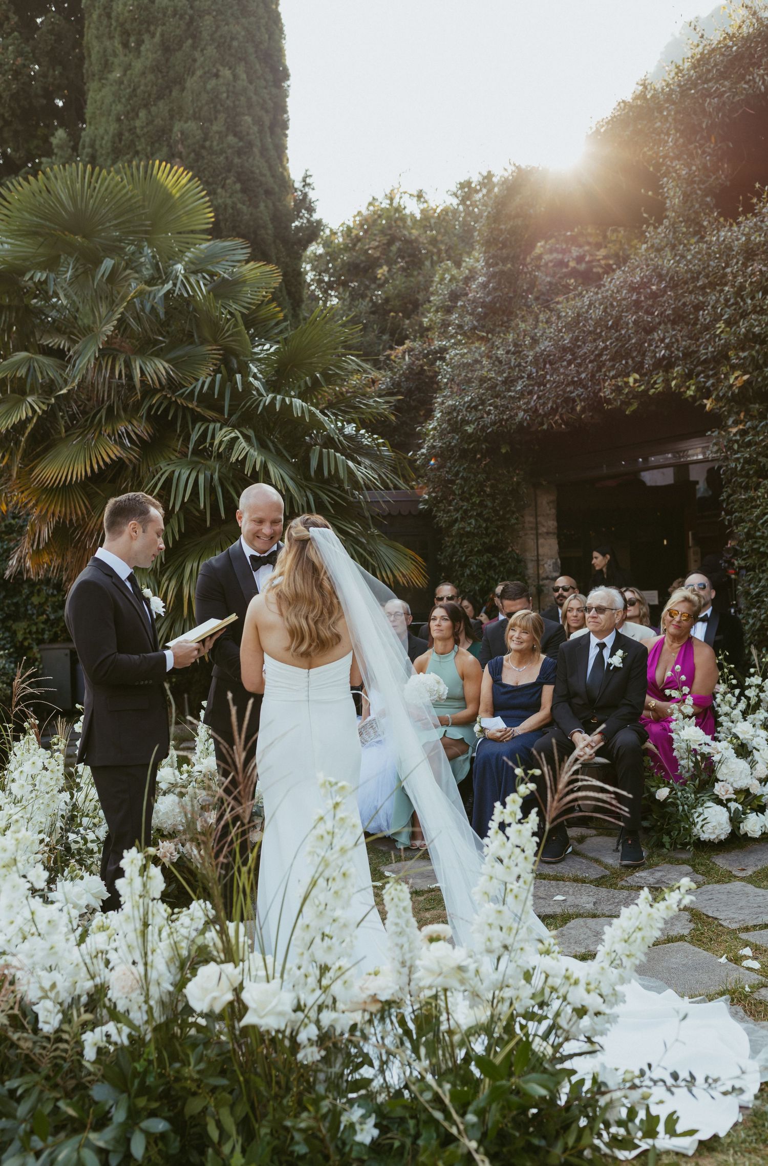 Bride and groom exchange vows outdoors surrounded by lush greenery and white flowers at a garden wedding ceremony.