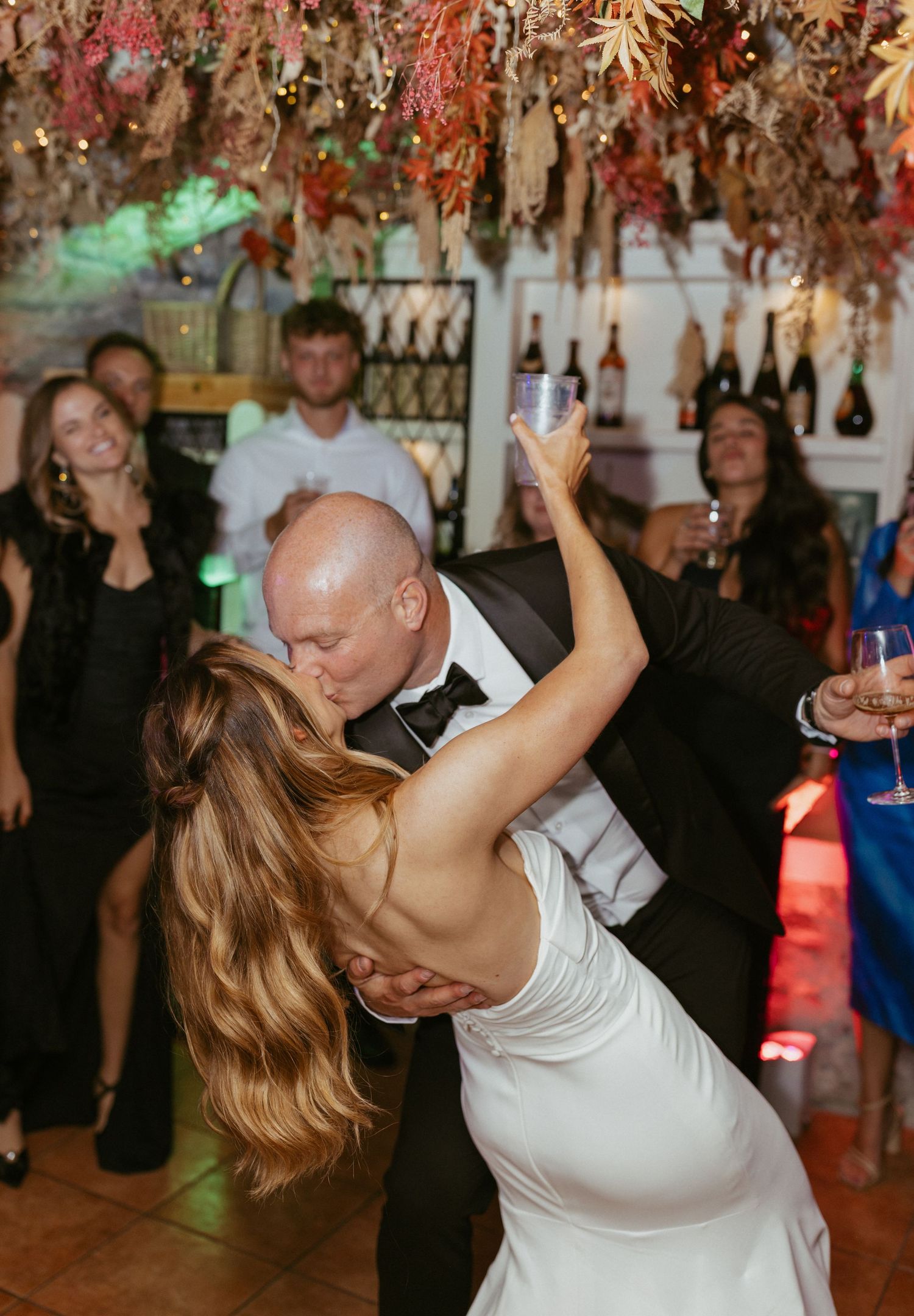 Groom dips bride for passionate kiss on dance floor surrounded by guests at wedding reception.