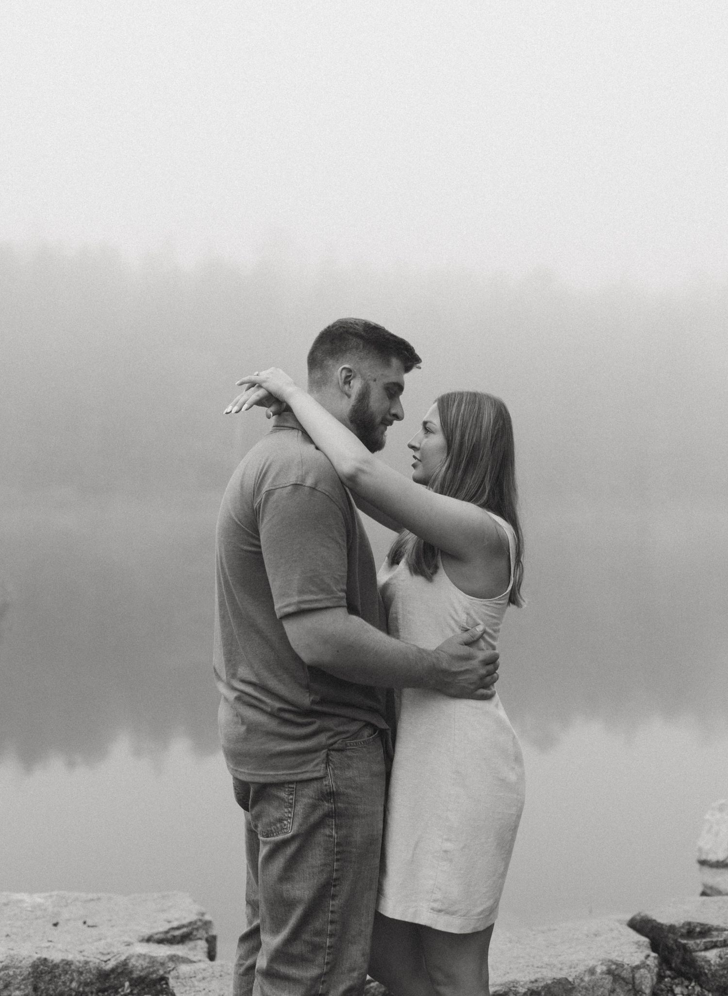 Black and white photo of a couple embracing tenderly by a foggy lakeside with reflections in the still water.