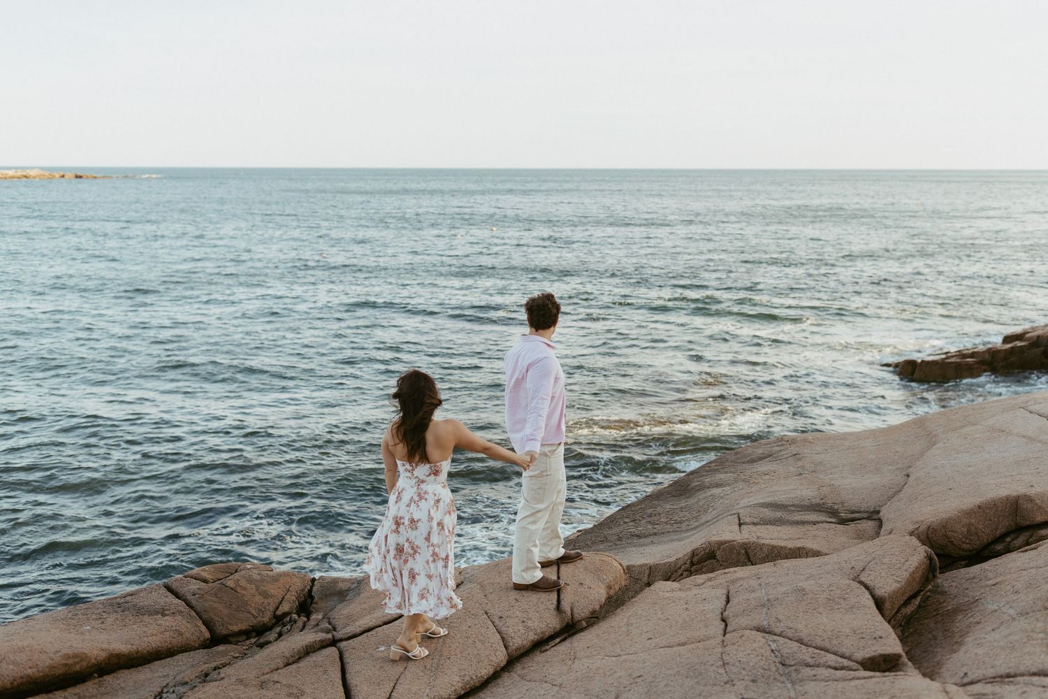 Couple holding hands while standing on rocky coastal Acadia cliffs overlooking the open ocean.