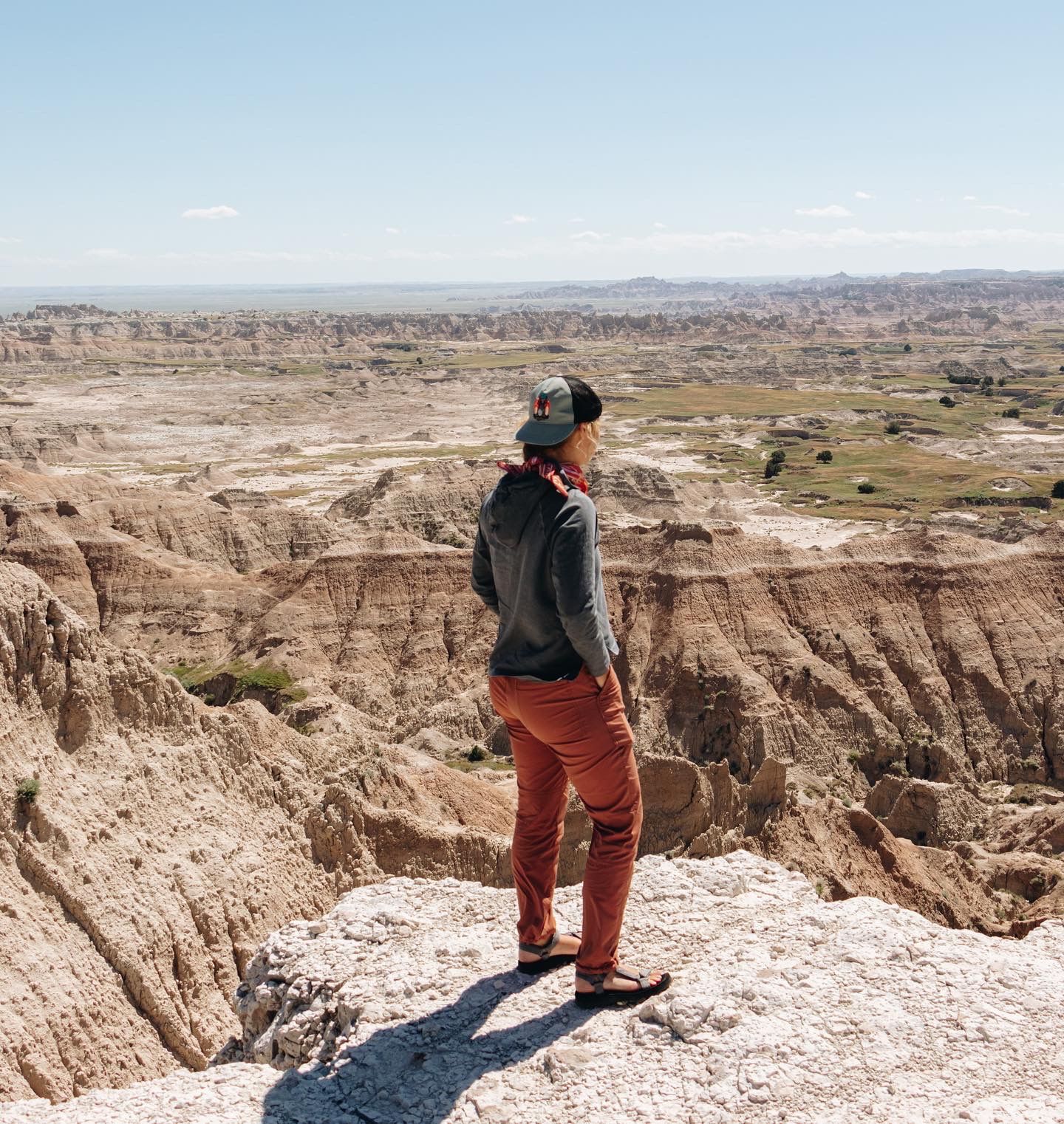 Man in red pants and cap stands on rocky overlook at Badlands National Park surveying vast eroded landscape.