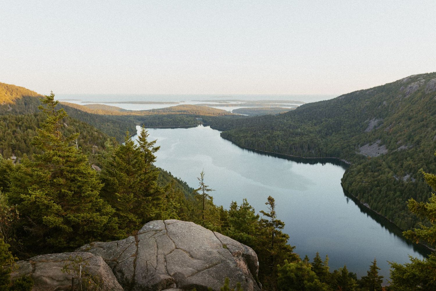 Aerial view of a serene lake surrounded by lush green forest and rocky cliffs under a clear sky.