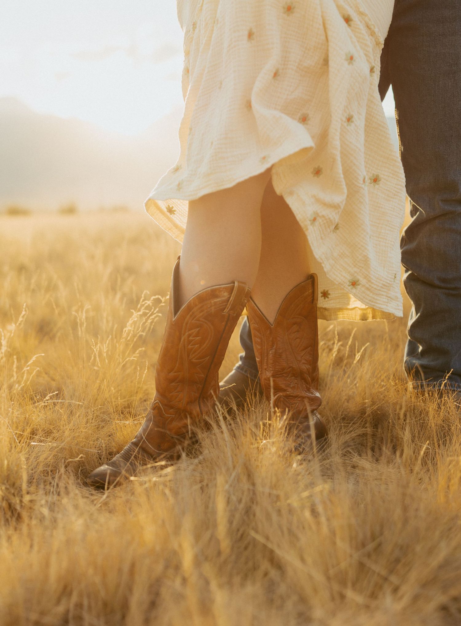 Close-up of bride wearing cowboy boots and floral dress standing in golden field at sunset.