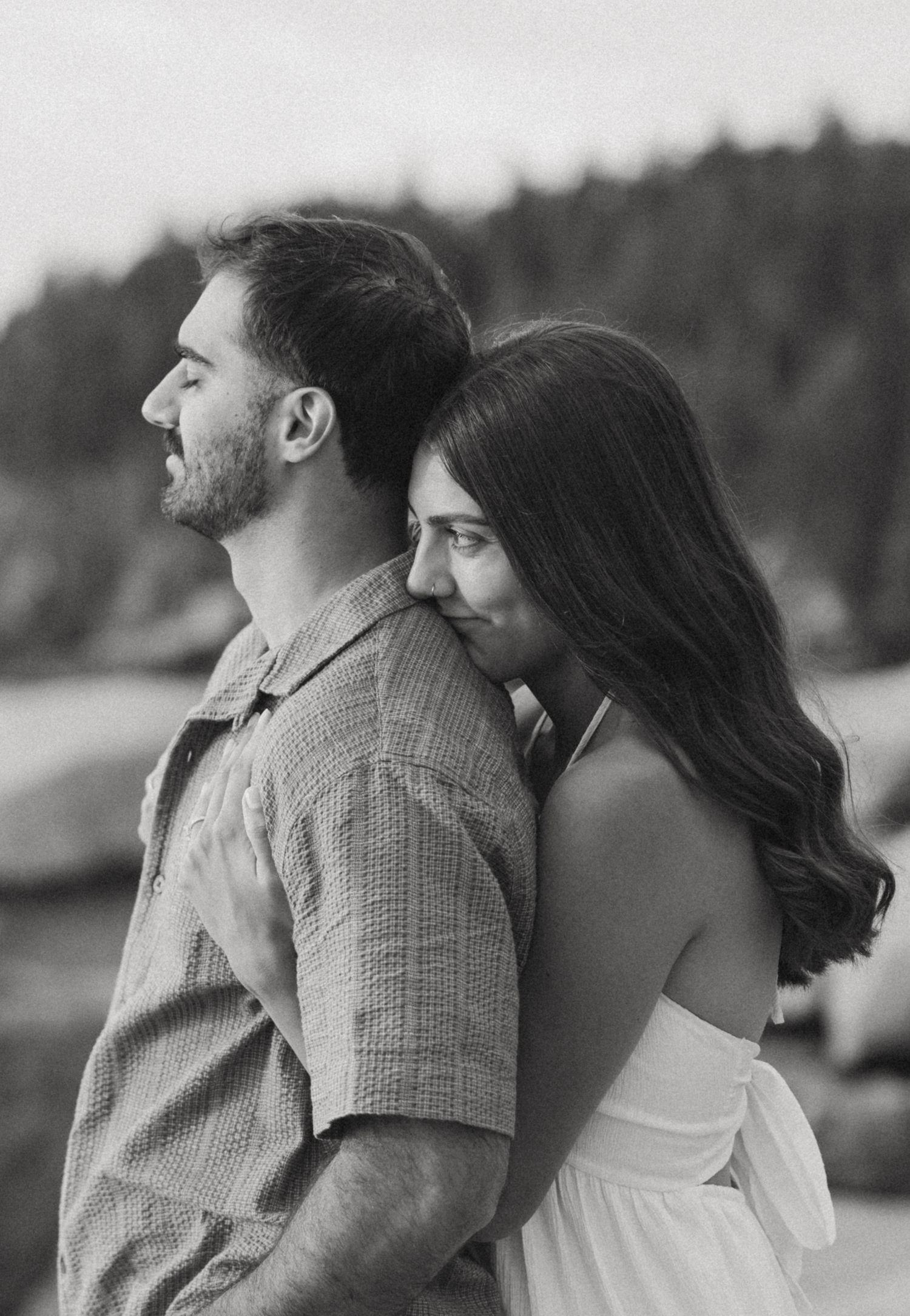 Black and white photo of a couple embracing outdoors, woman resting her head on man's shoulder near a forest.