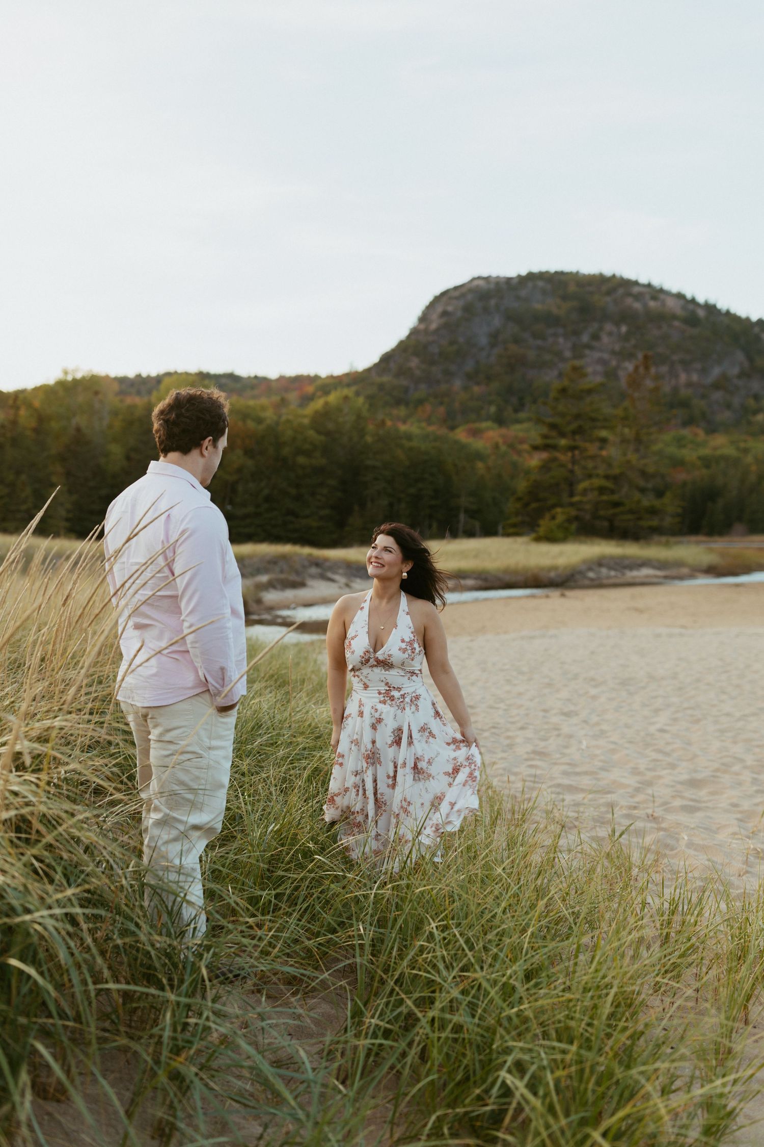 Couple walking through beach grass dunes with rocky Beehive Mountain and ocean backdrop.