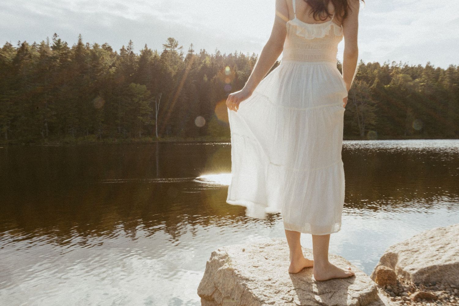 Woman in white dress standing barefoot on rocks by a sunlit lake surrounded by lush green forest.