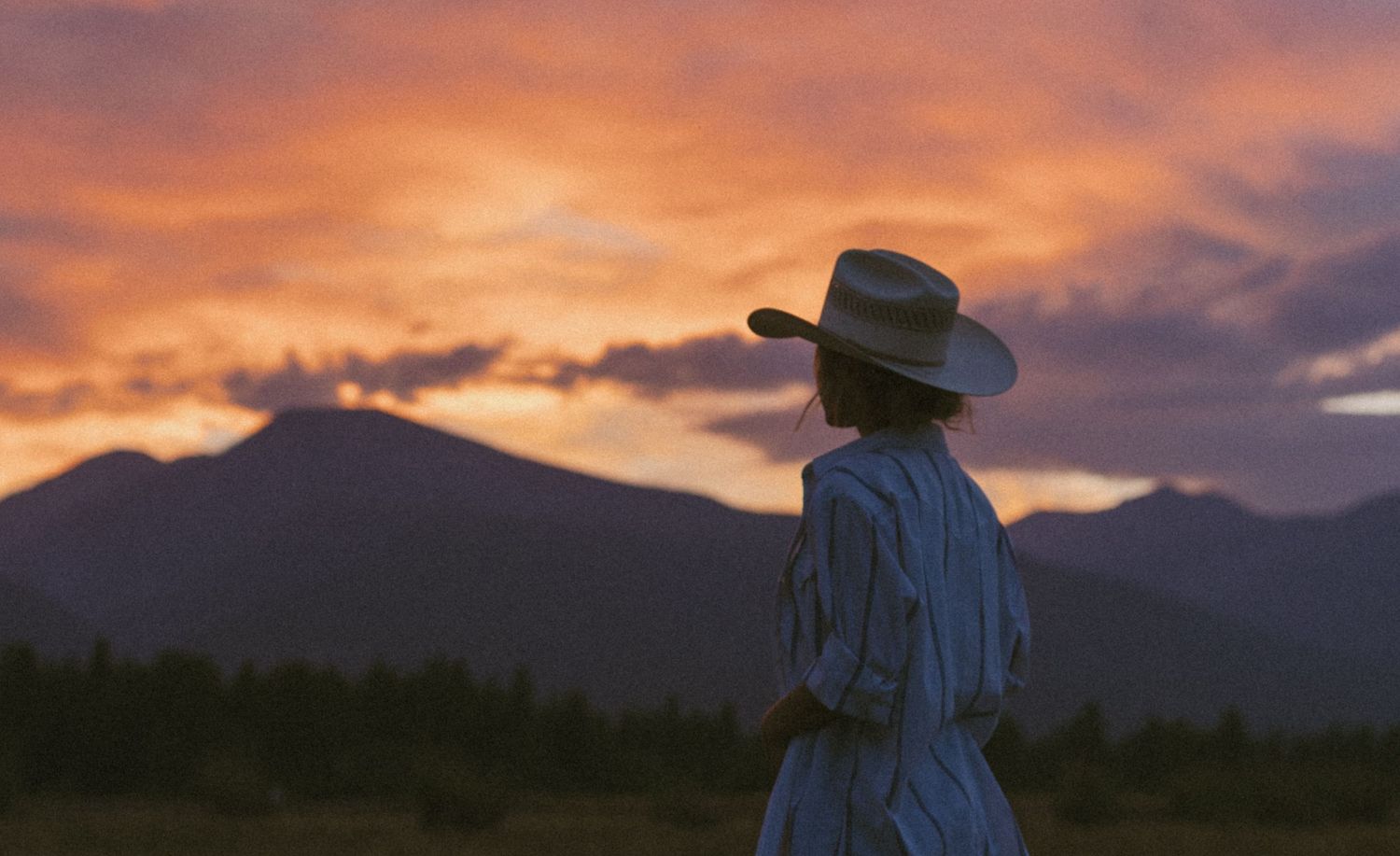 A woman in a cowboy hat and striped dress gazes at a dramatic orange sunset over mountain silhouettes.