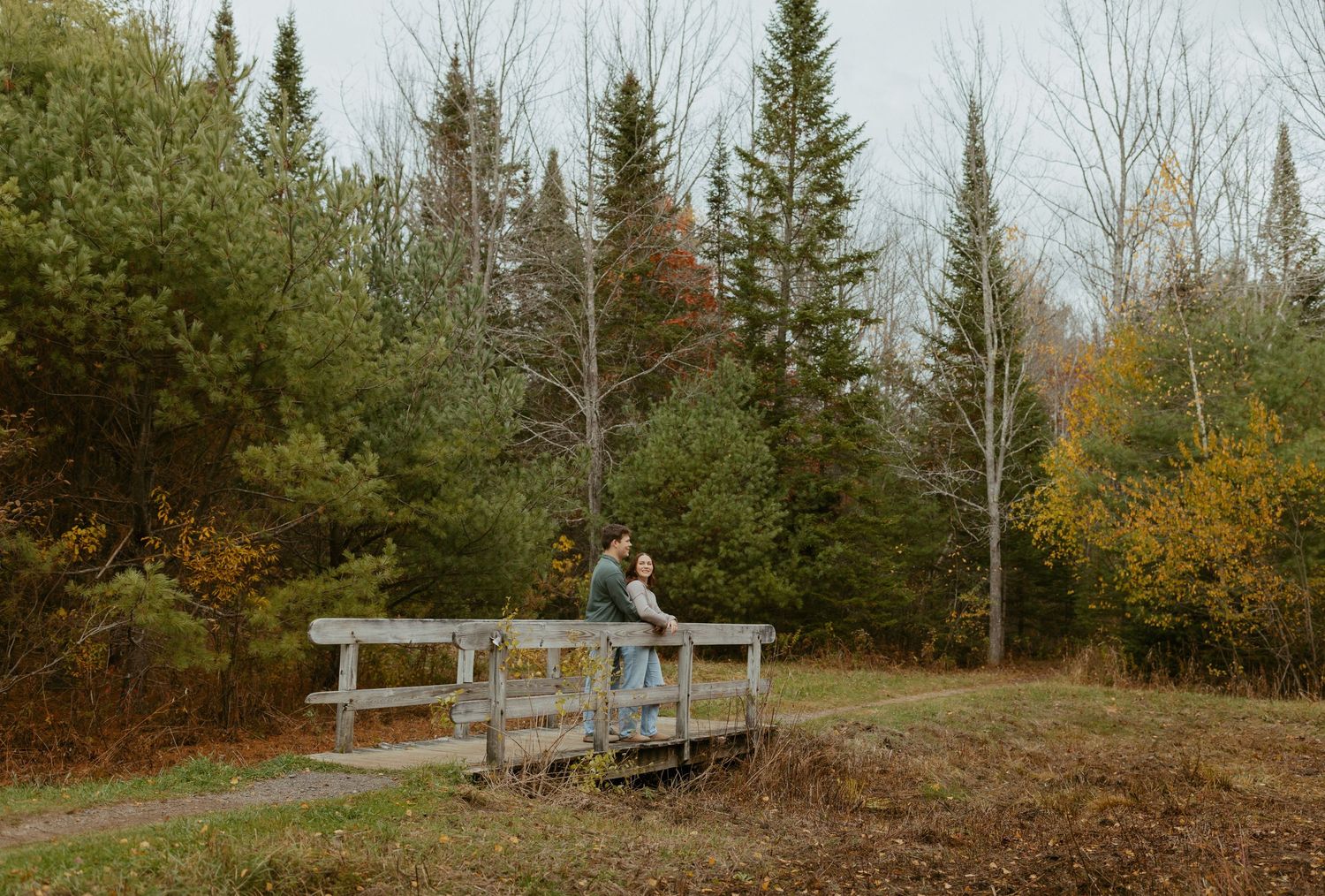 Couple cuddling together on a weathered wooden bridge amid lush fall landscape scenery.