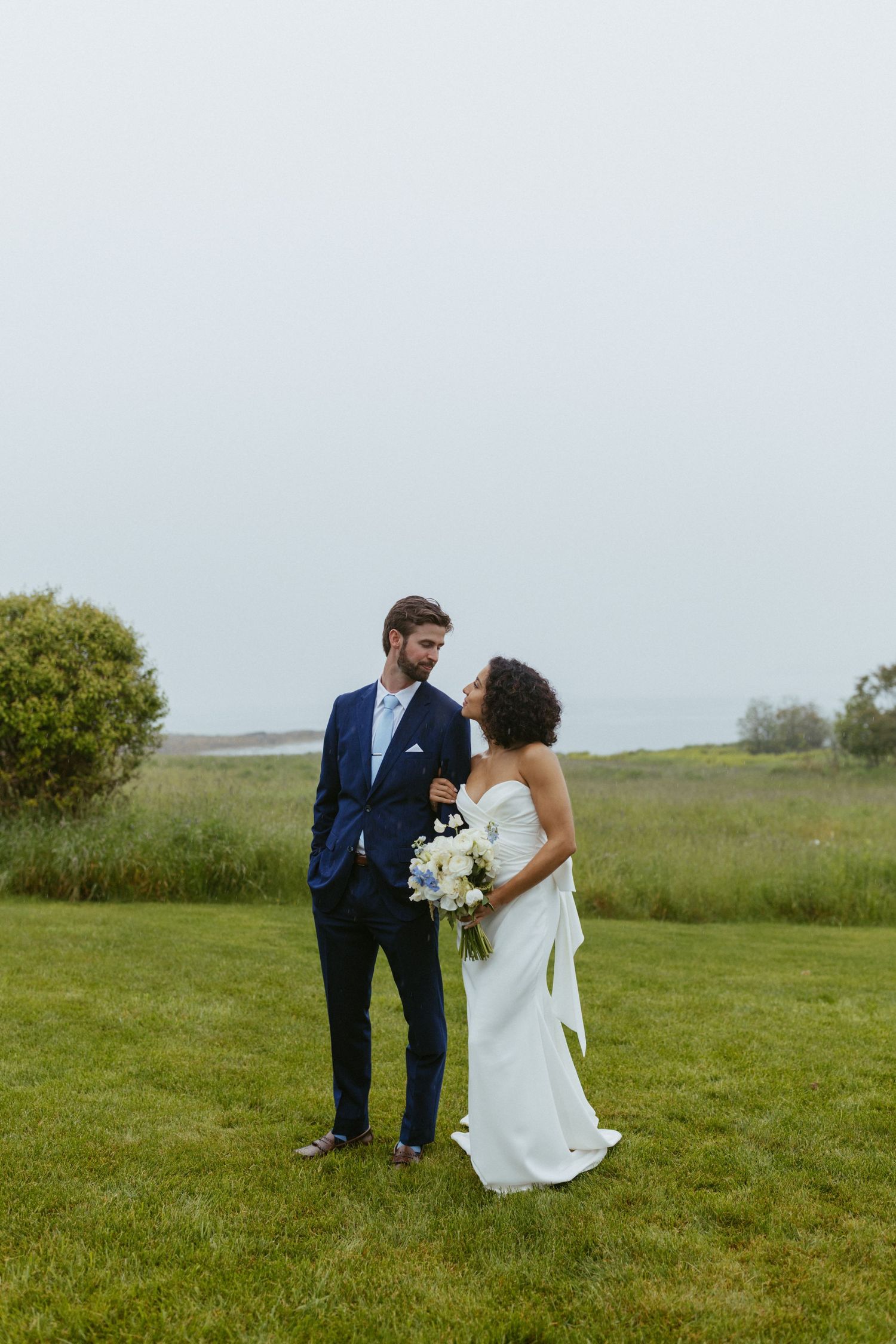 A bride in a white gown holding a bouquet stands with a groom in a navy suit on a green lawn by the Maine coast.