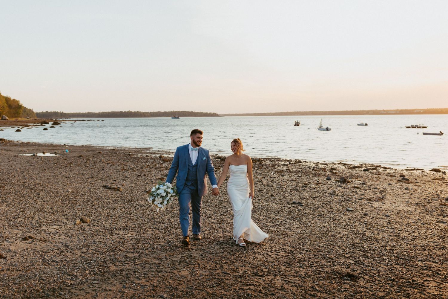 Bride and groom walk hand-in-hand along a rocky Maine beach at sunset near calm ocean waters.
