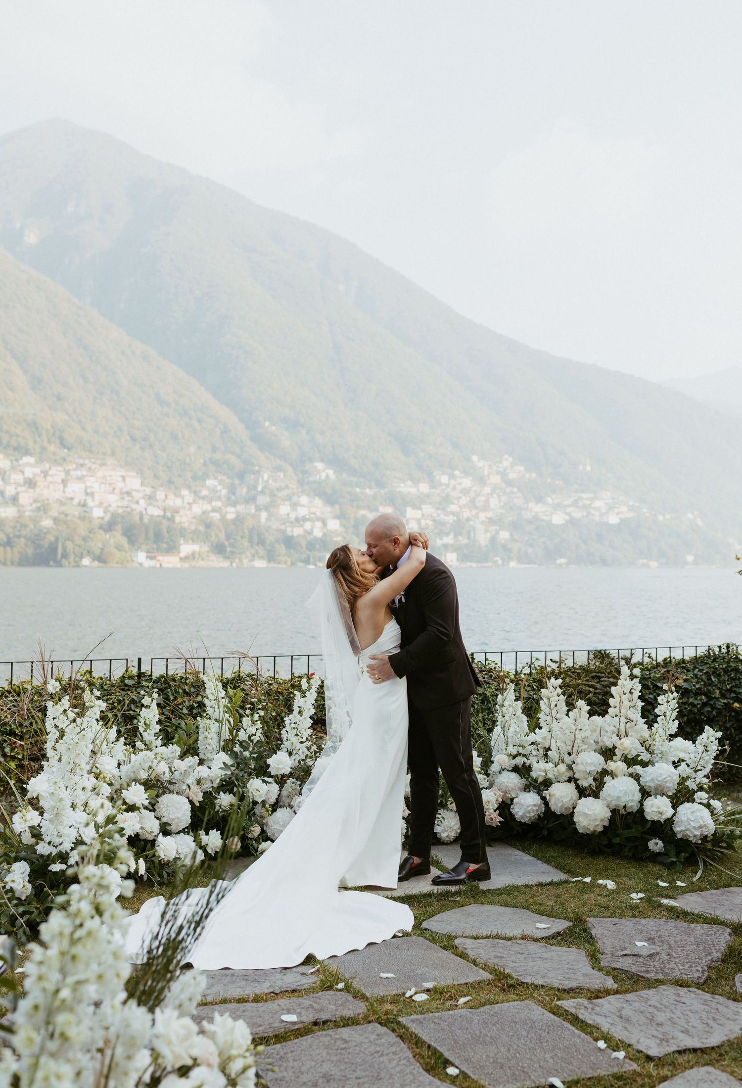 Bride and groom share a kiss at Lake Como wedding ceremony with white floral arrangements and mountain views.