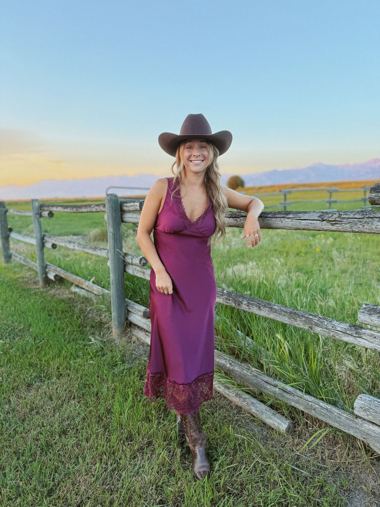 Woman in burgundy midi dress, cowboy hat, and boots leaning on wooden fence at sunset on a ranch.