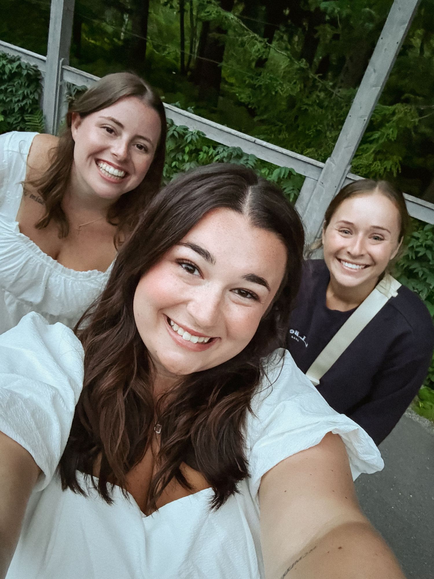 Three smiling young women taking a selfie outdoors near lush green trees and a glass greenhouse.
