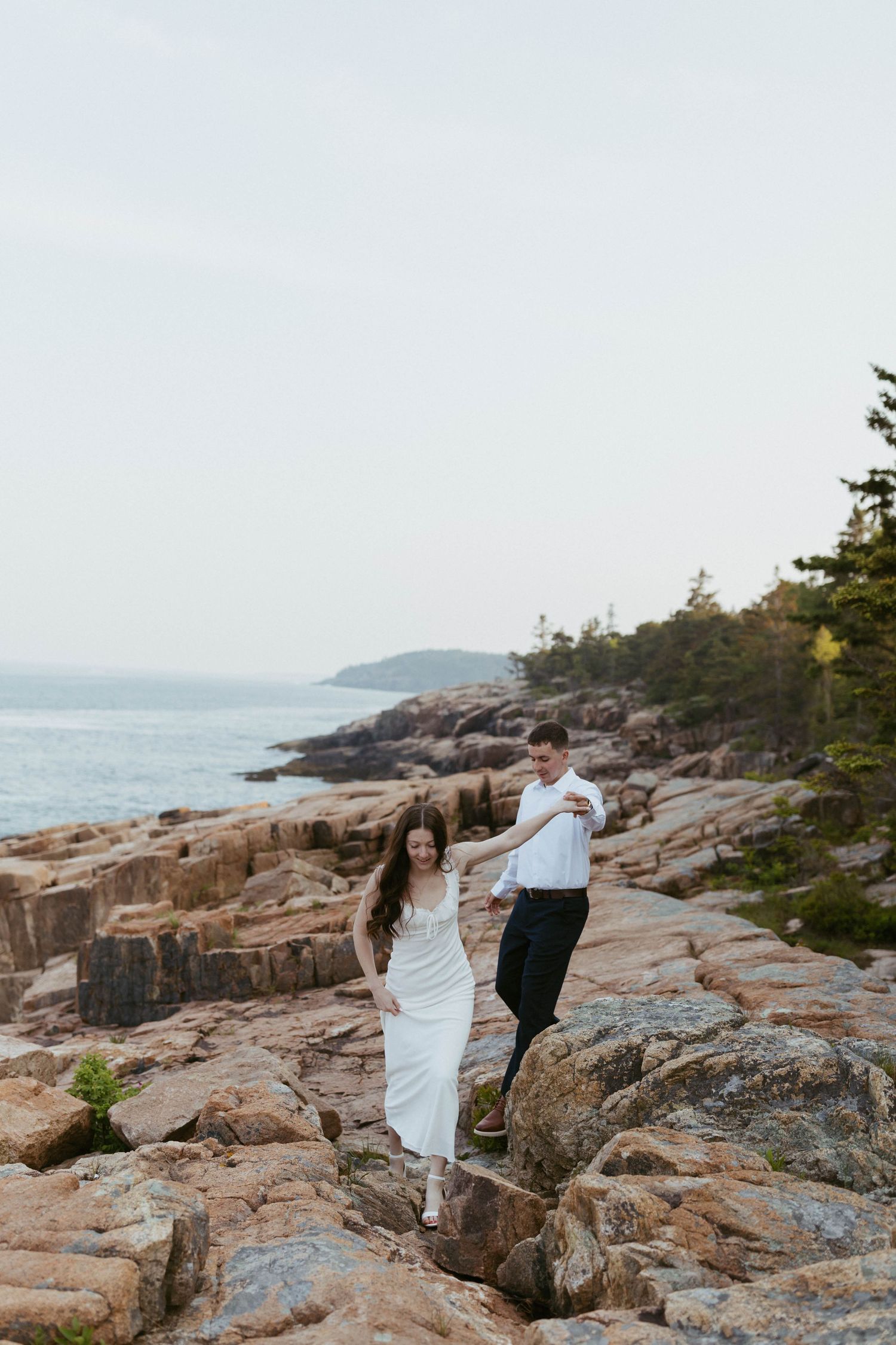Couple walking hand in hand across rocky coastal terrain with evergreen trees and ocean in background.