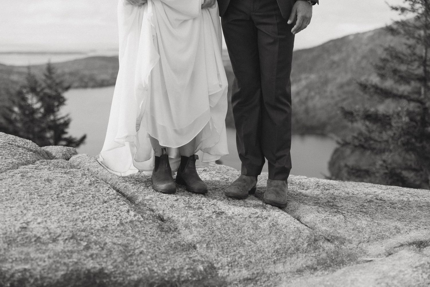 Black and white photo of bride and groom standing on rocky mountain summit wearing boots, with lake and trees in background.