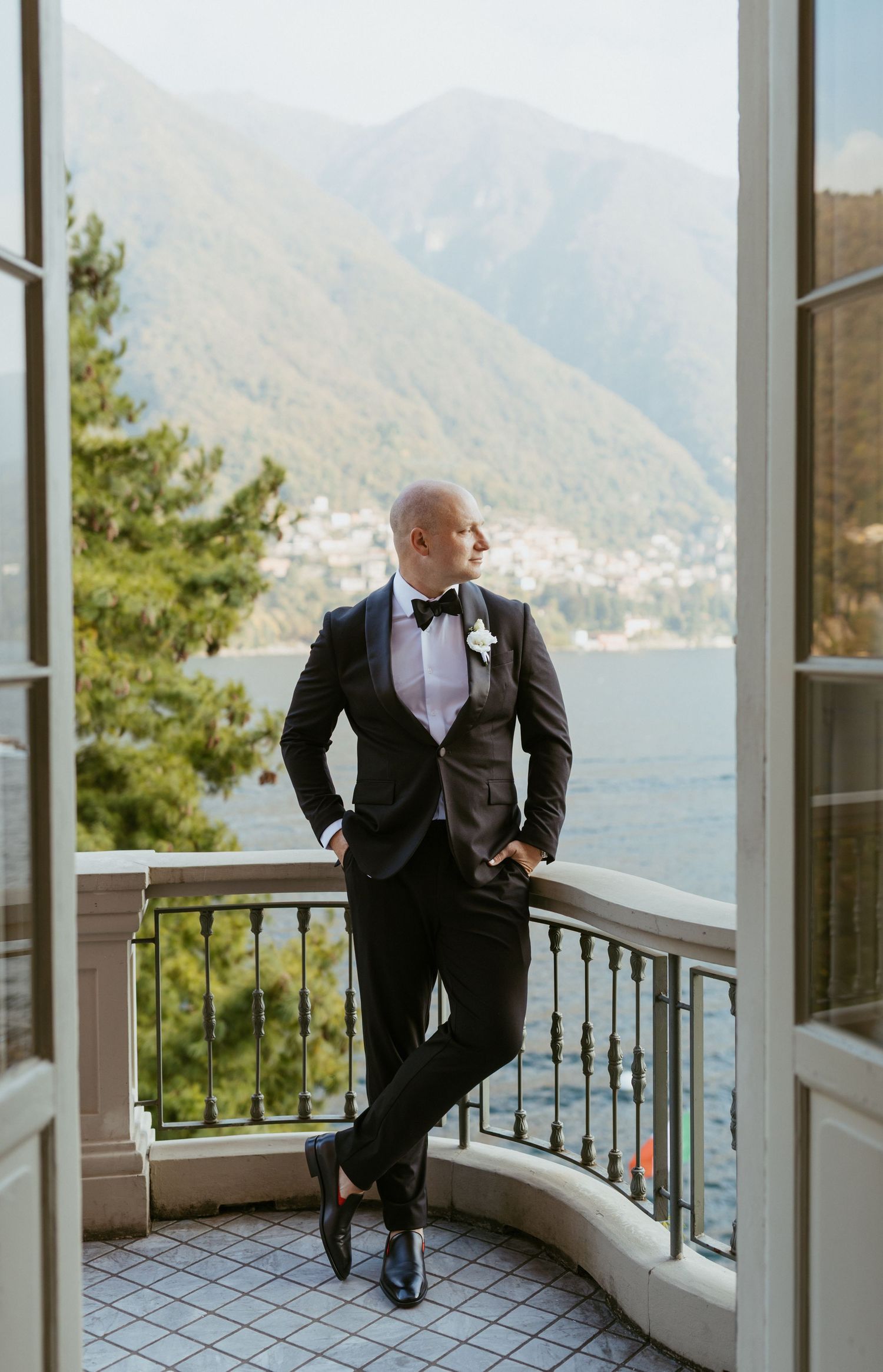 Groom in black tuxedo with bow tie stands on balcony overlooking lake and mountains at elegant wedding venue.